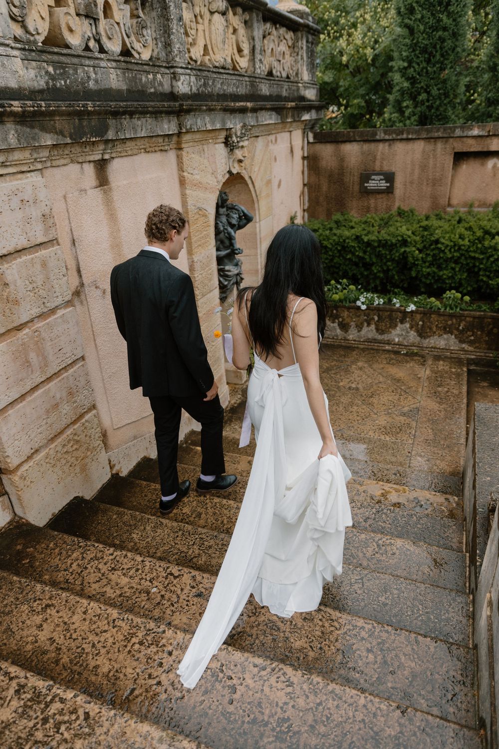 A couple descends stone steps in formal attire against an ornate garden wall with sculptural details.
