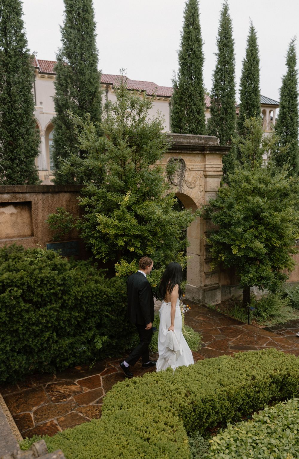 A peaceful garden path lined with tall cypress trees and lush greenery surrounds a Mediterranean-style estate.