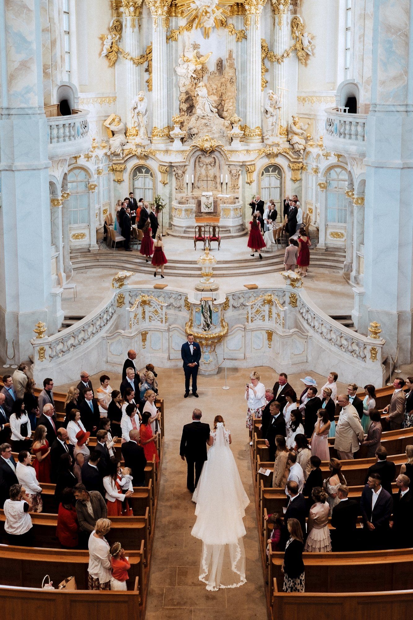 Wedding ceremony in an ornate Baroque church with gold details and white walls as guests observe from wooden pews.