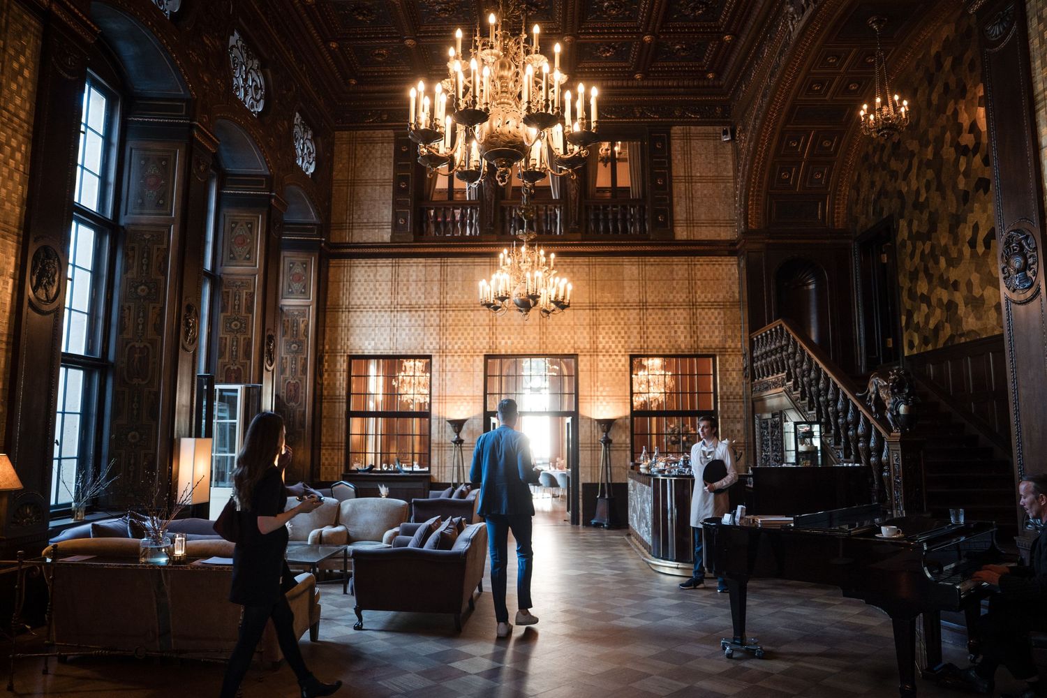 A silhouette stands in an elegant vintage room with crystal chandeliers, grand piano and ornate woodwork details.