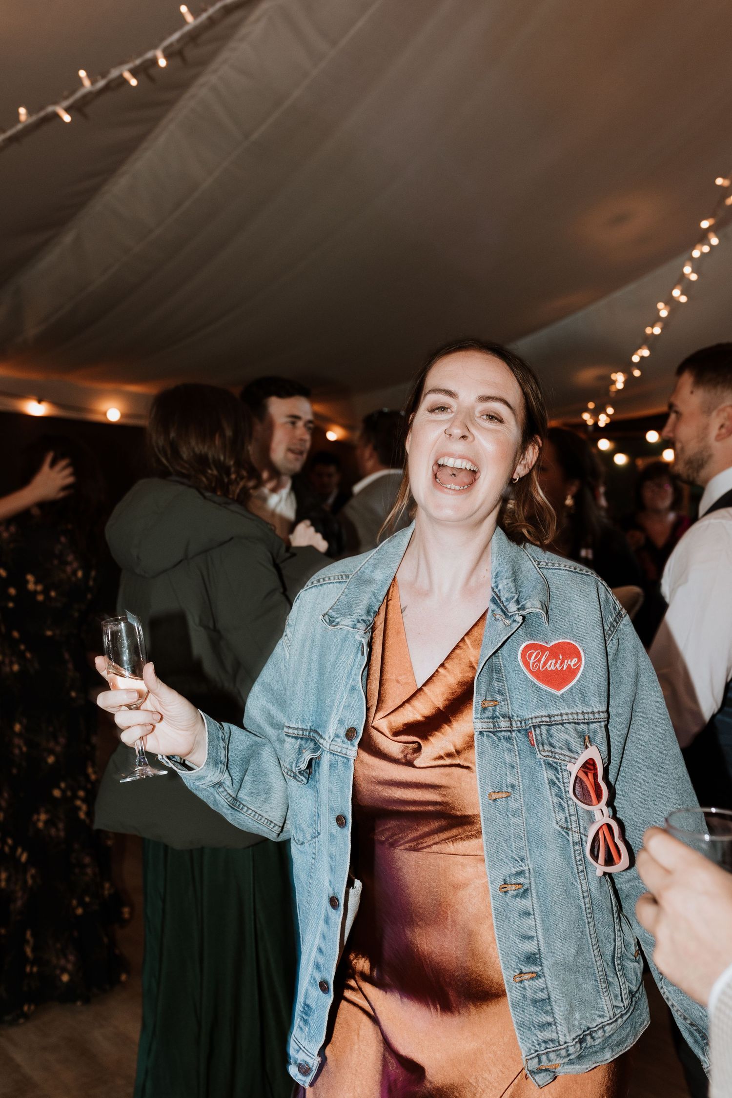 Person wearing denim jacket with heart patch and orange dress dances with champagne glass at indoor party with twinkle lights.