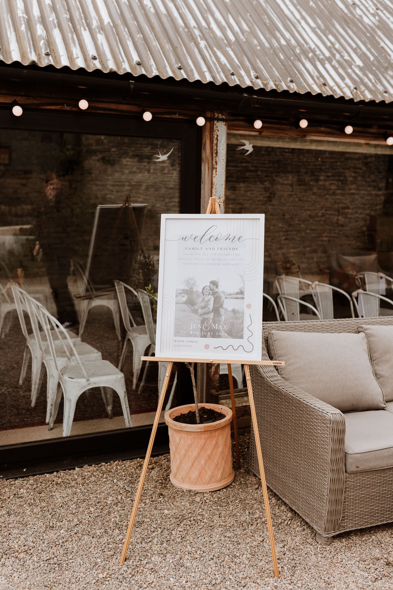 A rustic wedding welcome sign on a wooden easel stands beside rattan furniture and string lights under a covered patio.