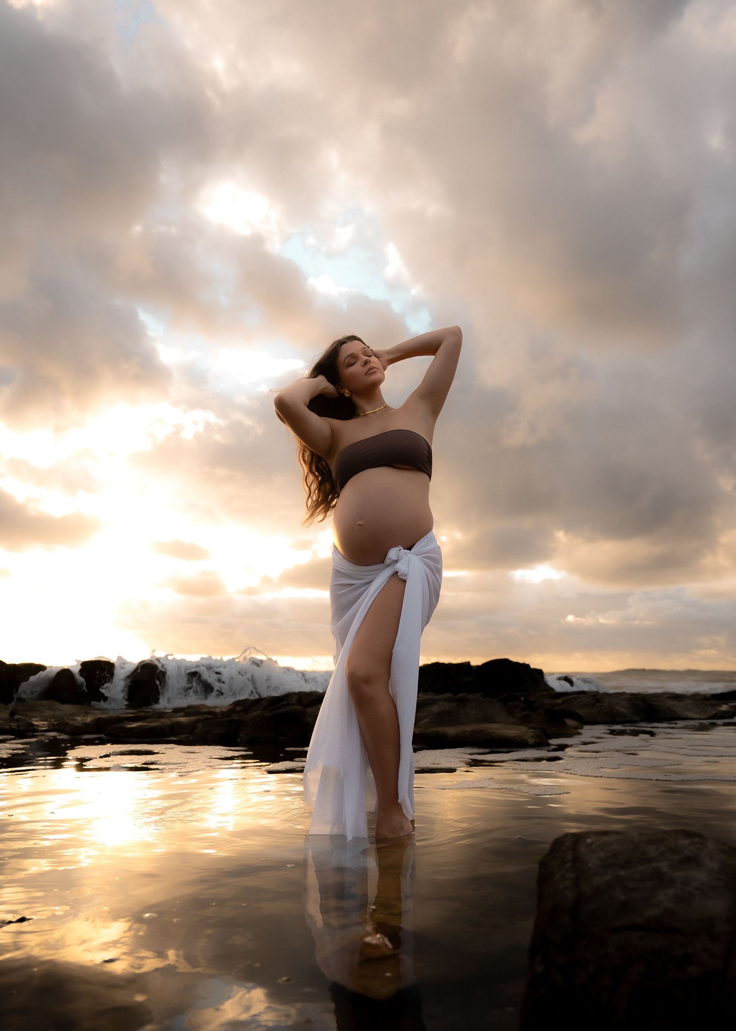 A dramatic maternity photo at sunset with a woman standing in shallow water among rocks.