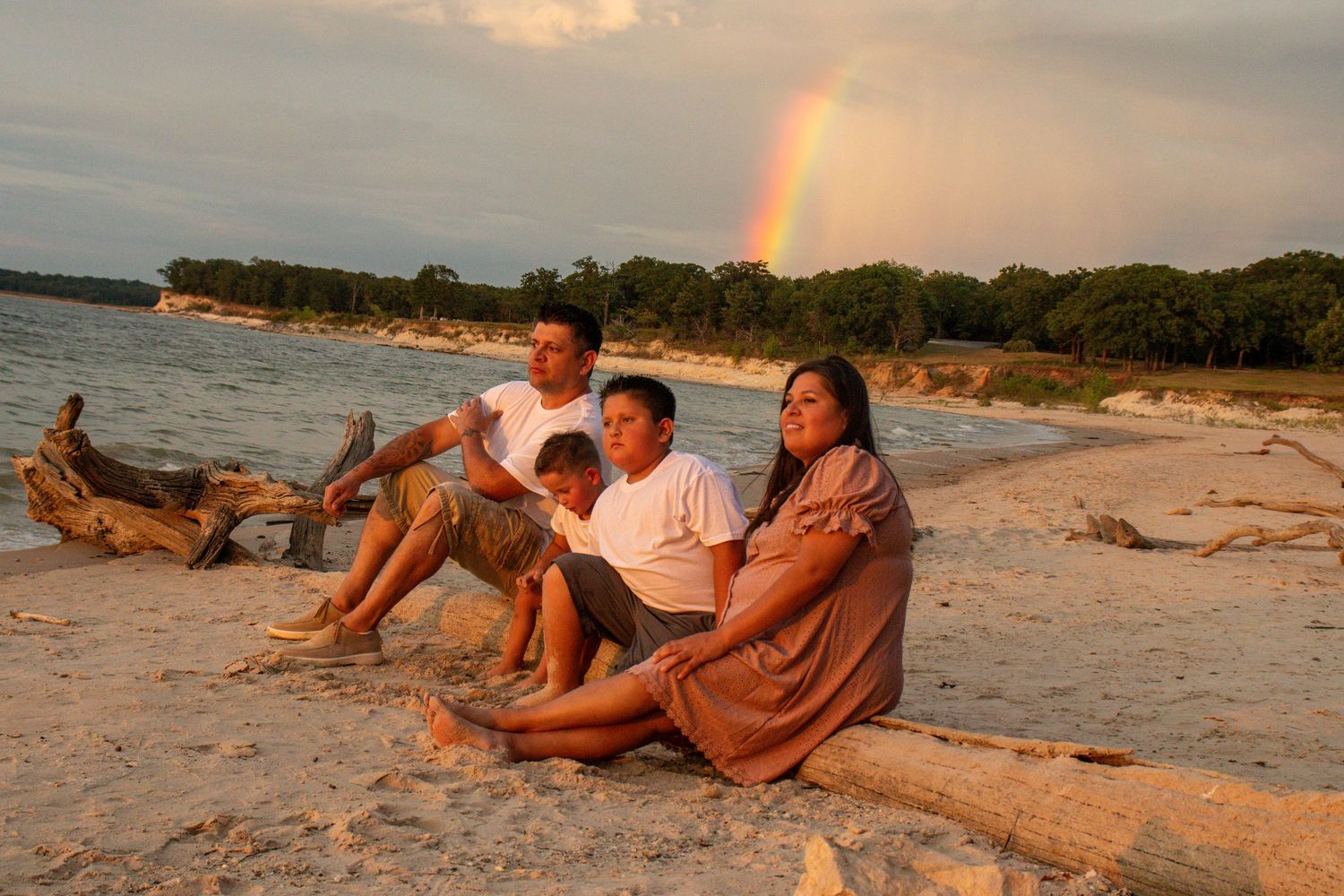 People relaxing on a sandy beach at sunset with a rainbow visible in the cloudy sky.