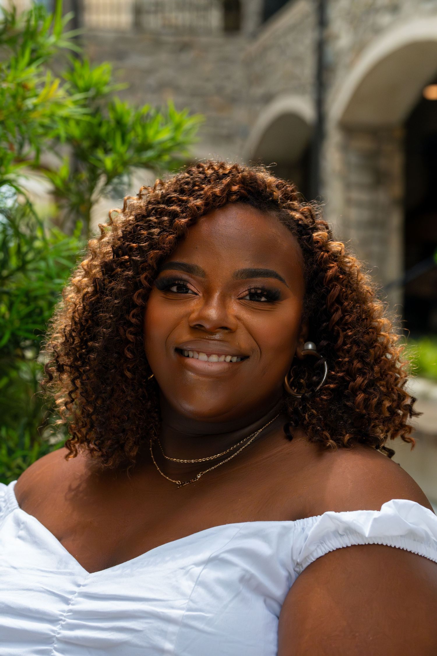 A photograph showing a bright smile and curly brown hair wearing a white off-shoulder top outside a stone building.