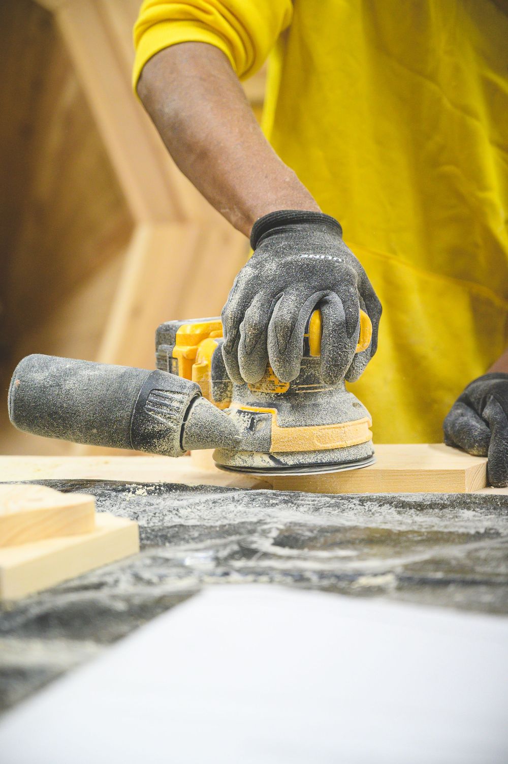 Person wearing yellow shirt using orbital sander on wood surface with work gloves and sawdust visible.