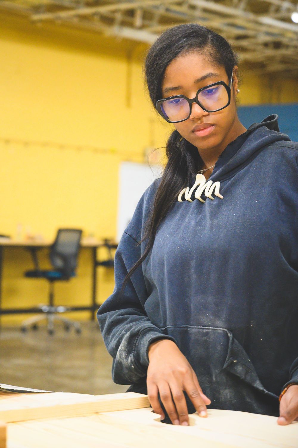 Student wearing hoodie and glasses in a classroom with yellow walls.