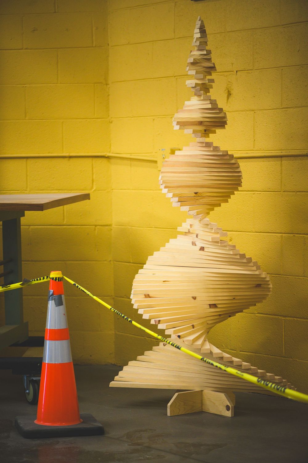 Artistic wooden spiral sculpture with orange traffic cone and caution tape against yellow wall.