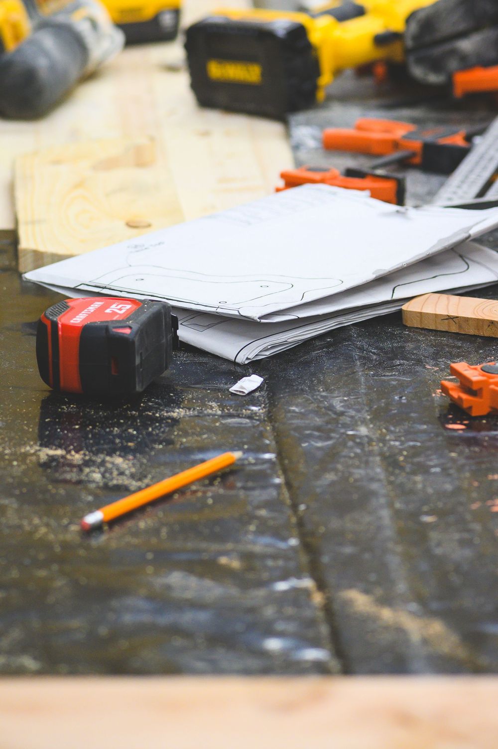 Work tools and papers scattered on a dark textured surface with yellow power tools visible in the background.
