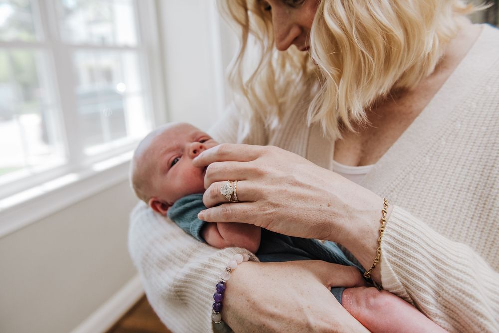 A parent gently bottle feeds an infant while sitting near a window in a cream-colored sweater.