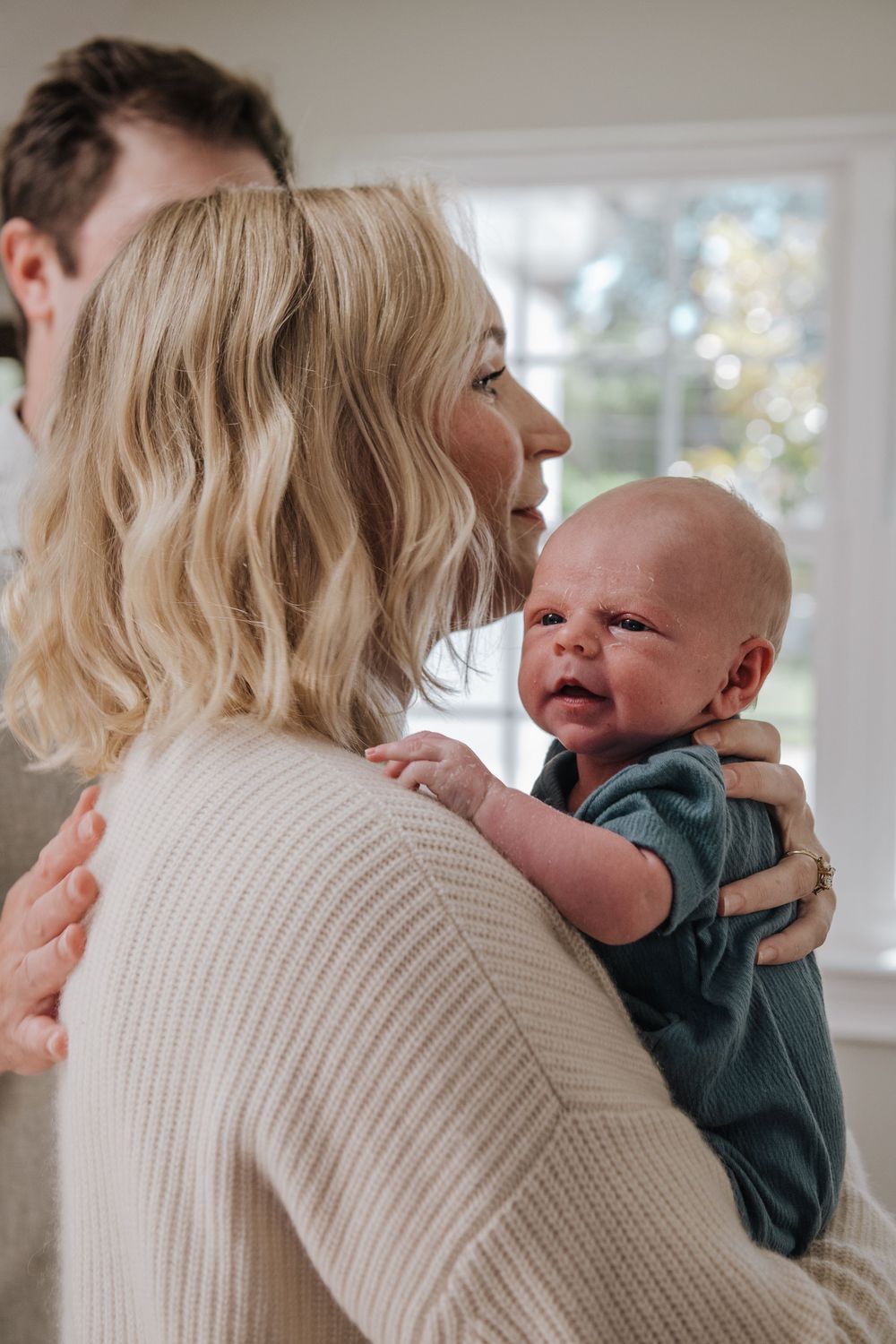 A baby in a dark green outfit smiles while being held by someone wearing a cream colored knit sweater.