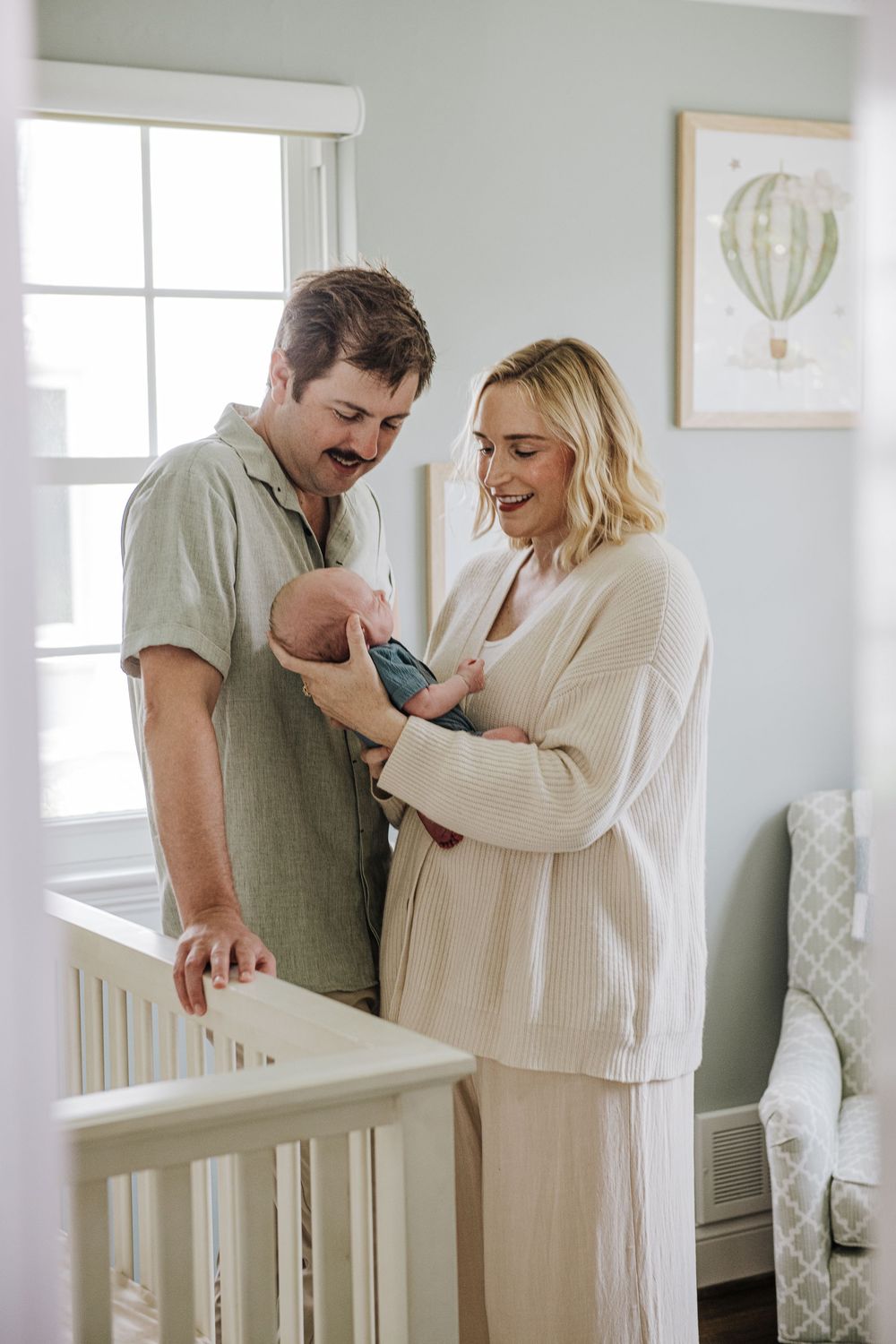A couple stands together by a white crib in a nursery with mint green walls and decorative hot air balloon artwork.