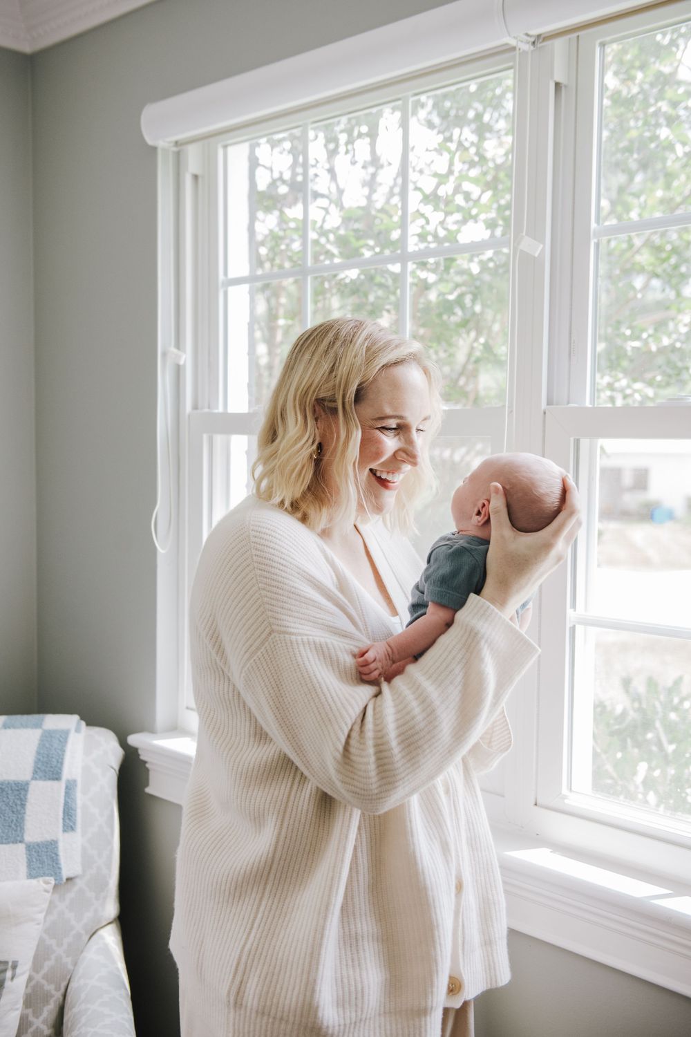 A person wearing a cozy white cardigan holds a newborn baby while standing by bright windows in a nursery.
