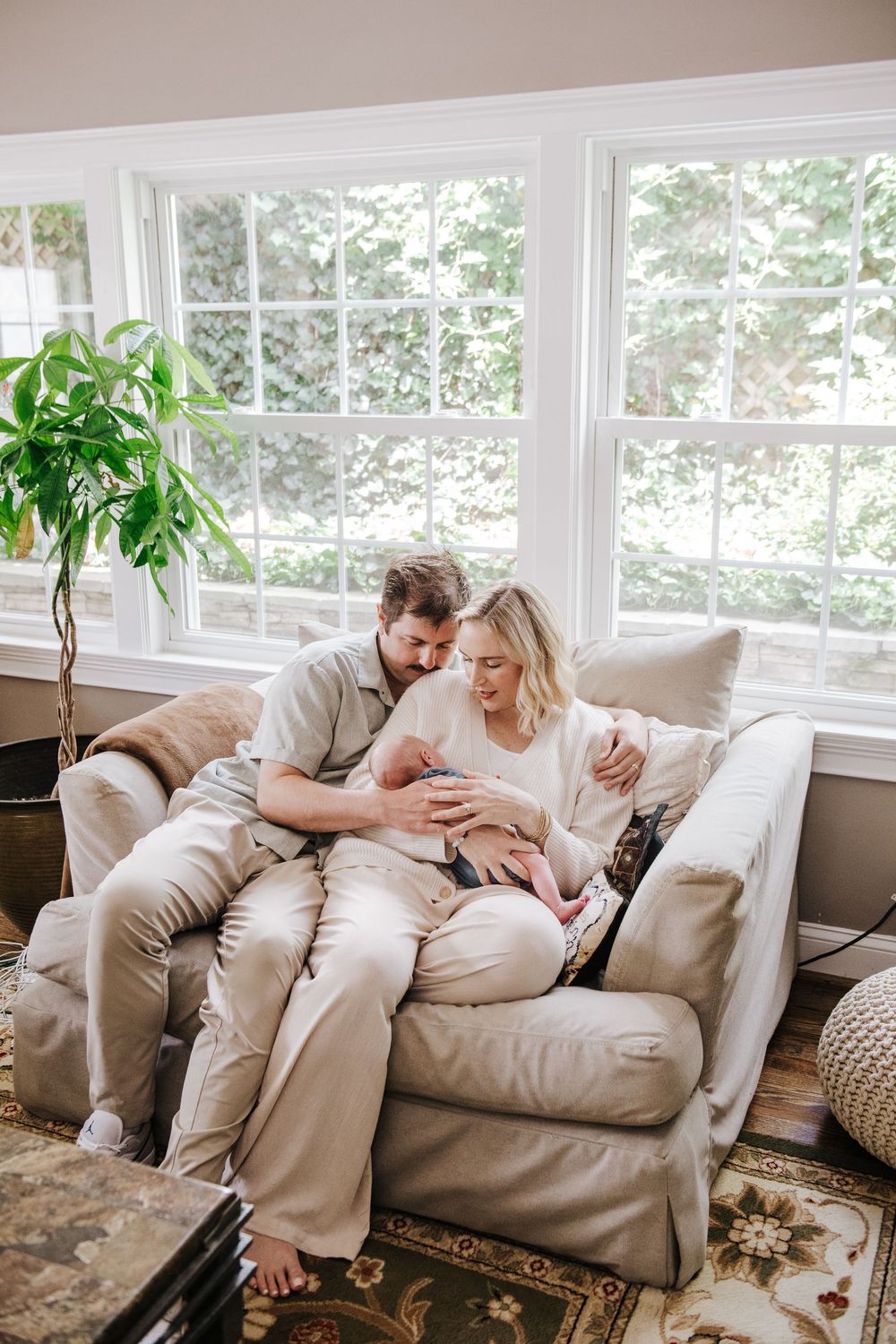 Couple relaxes comfortably together on a plush beige loveseat by bright windows in a sunny living room.