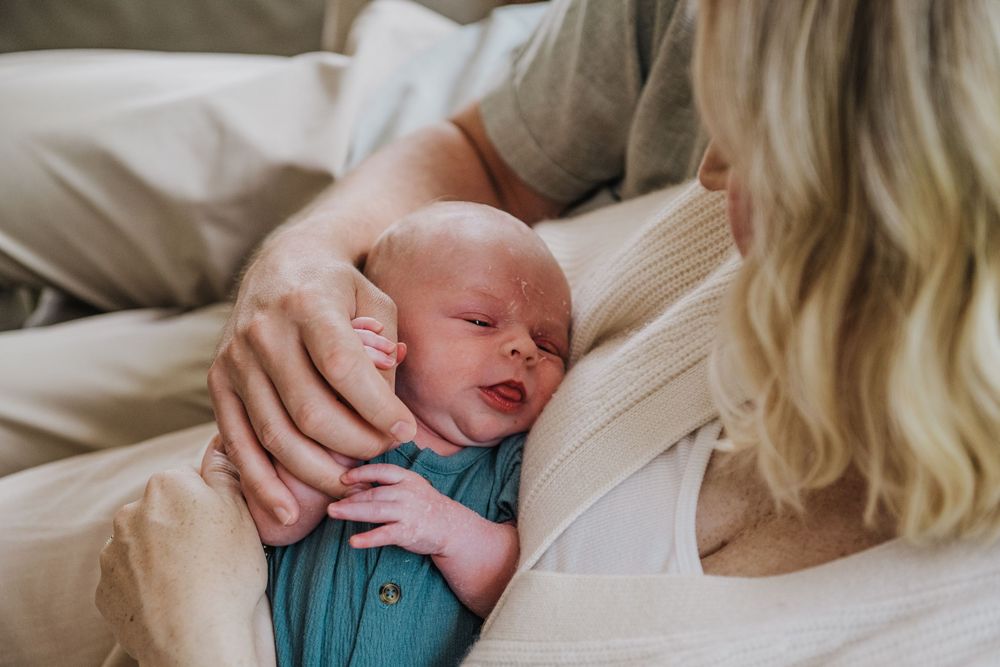 A newborn baby in a blue outfit sleeps peacefully in a caregiver's arms on white bedding.
