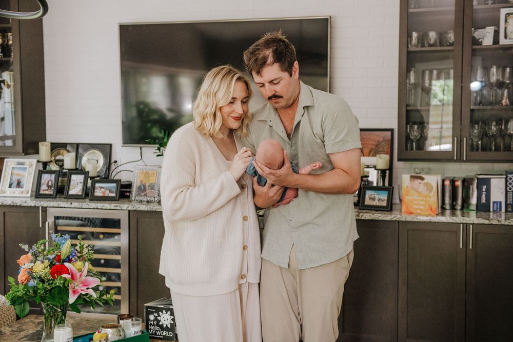 A couple stands together in a kitchen holding their newborn baby while wearing neutral colored casual clothing.