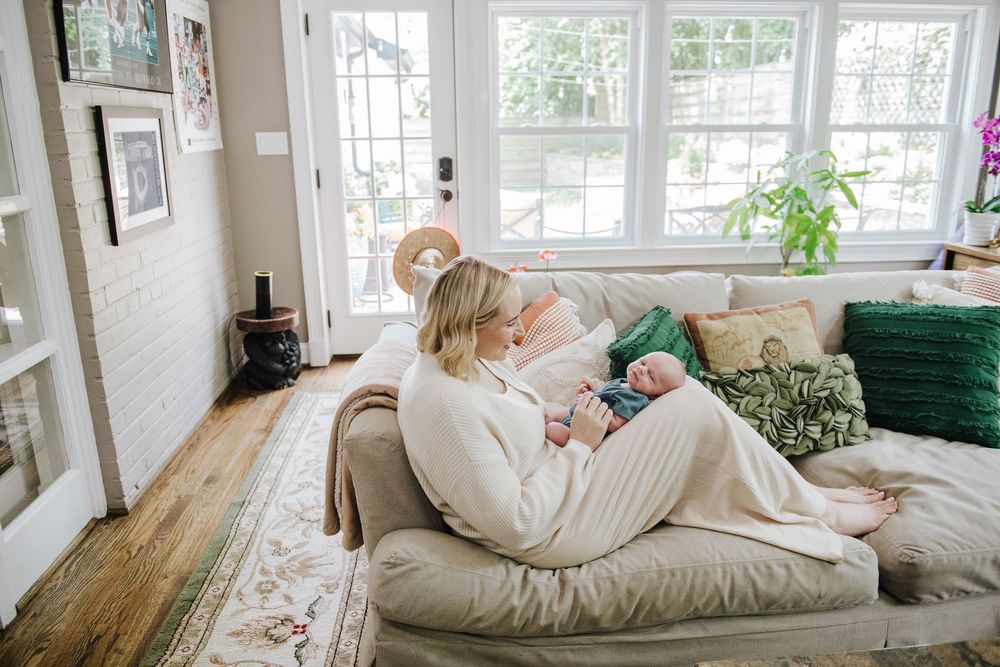A cozy living room scene with someone lounging peacefully on a beige couch near bright windows and green plants.