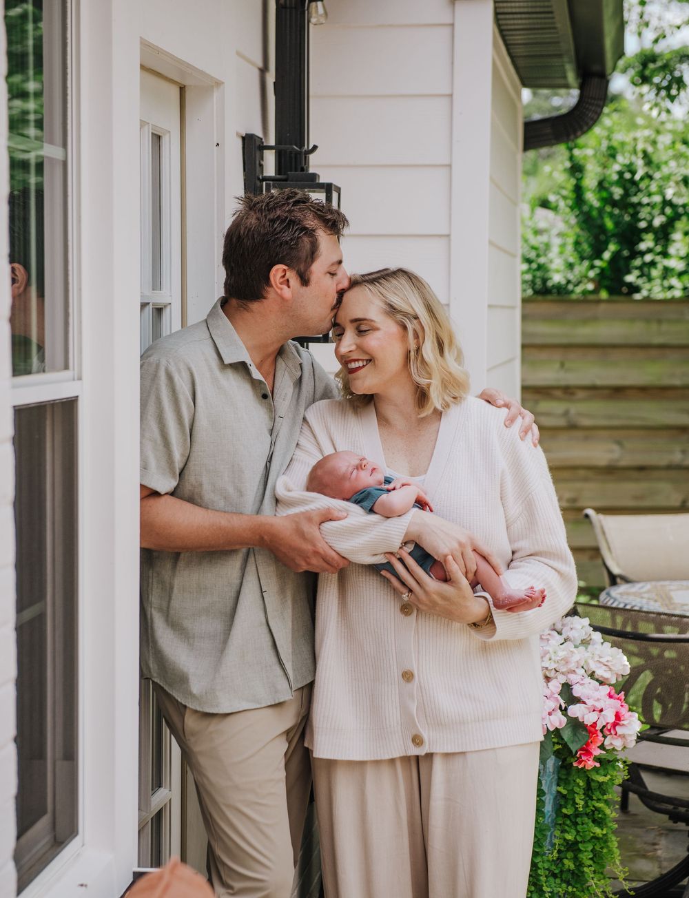 A couple stands lovingly on their porch holding a newborn baby wrapped in pink, surrounded by flowers and greenery.