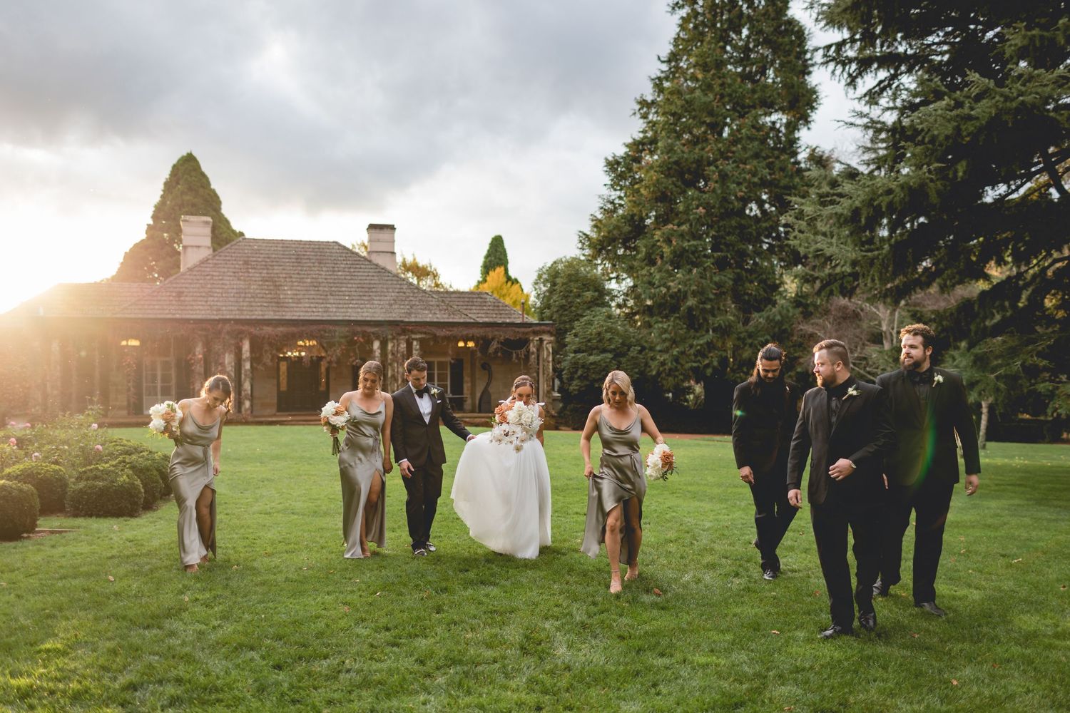 Wedding party walking and laughing together on a green lawn with sunlight and historic building in background.