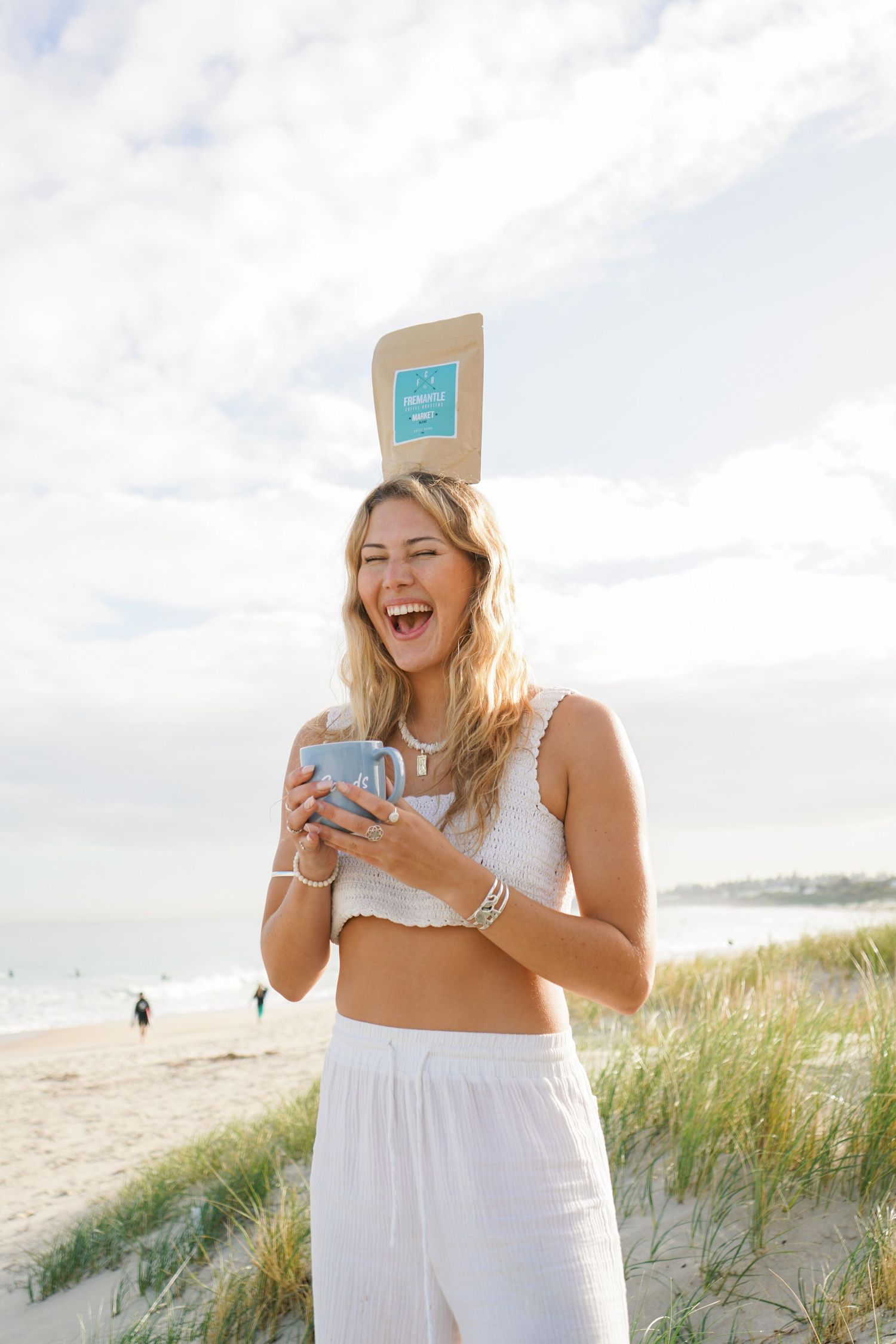 Woman in white outfit laughing while holding phone on sandy beach.