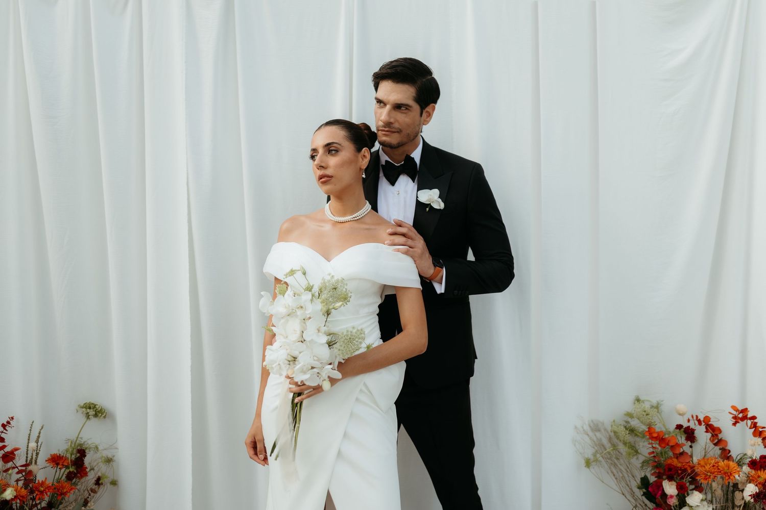 A bride in an white gown and groom in black tuxedo pose against a white backdrop with red floral accents.
Kima Tulum wedding photographer
