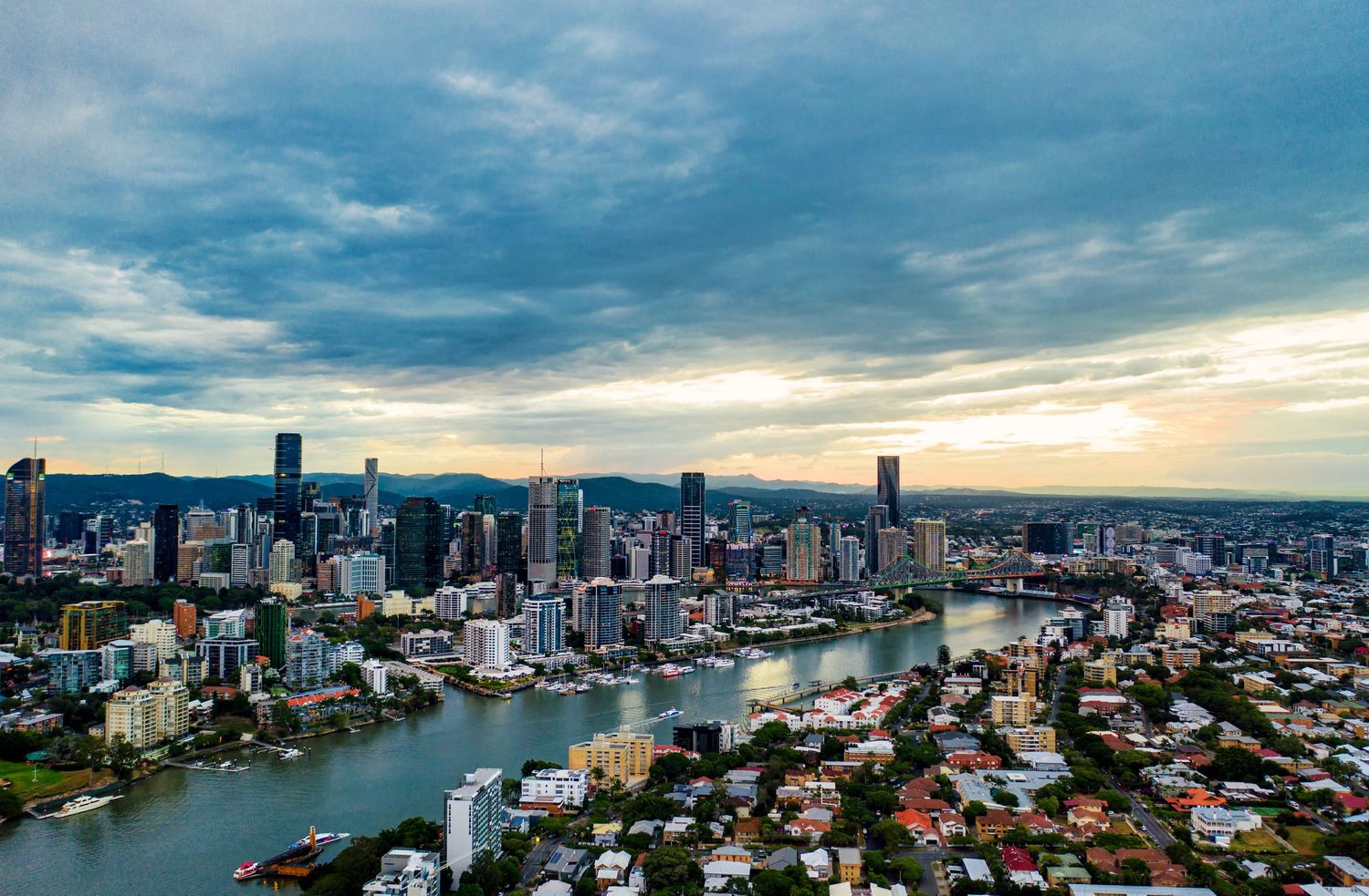 Brisbane cityscape with winding river, modern buildings and residential areas under cloudy sky.