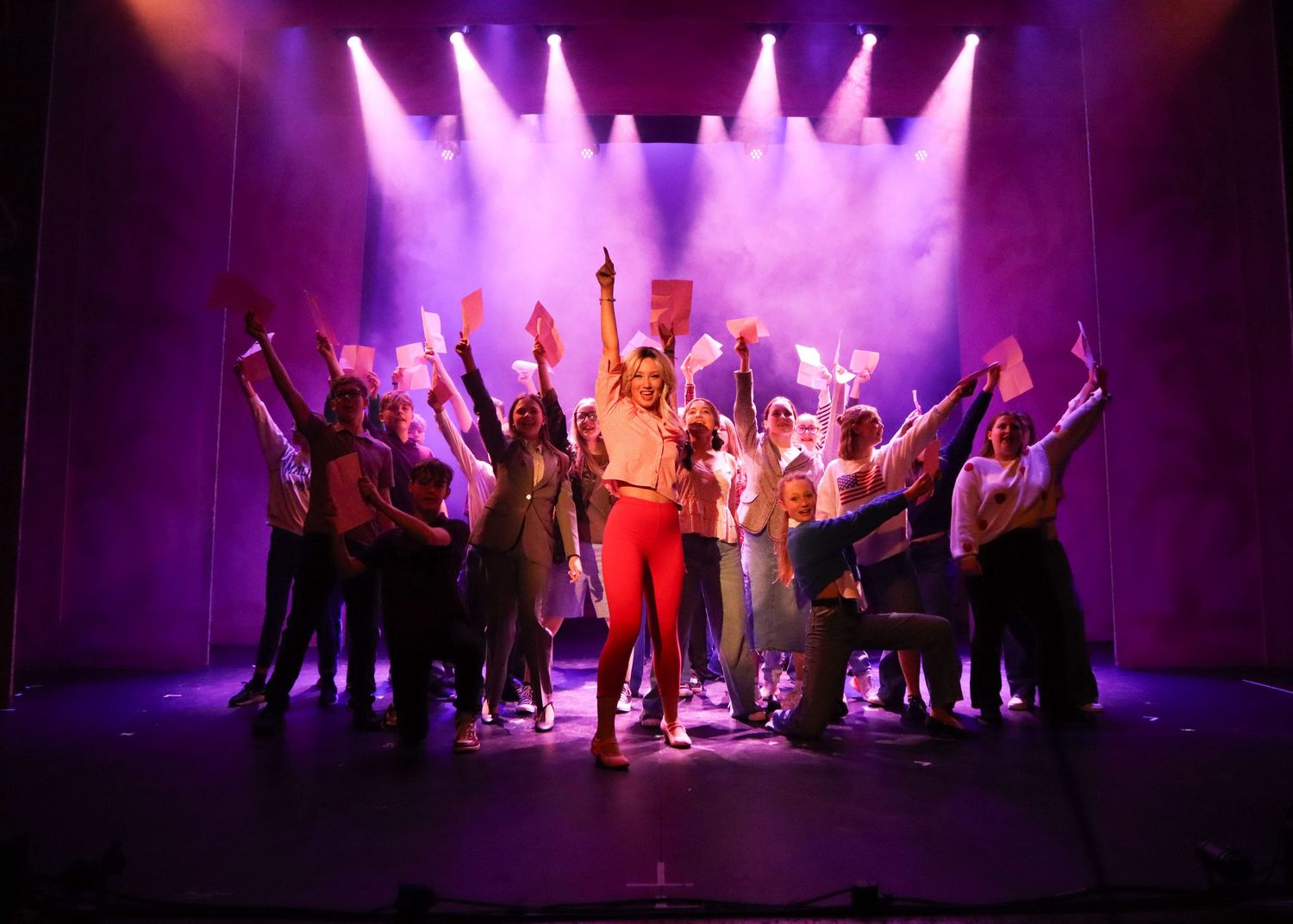 Performers pose dramatically on a purple-lit stage during a theatrical production with dynamic lighting effects.