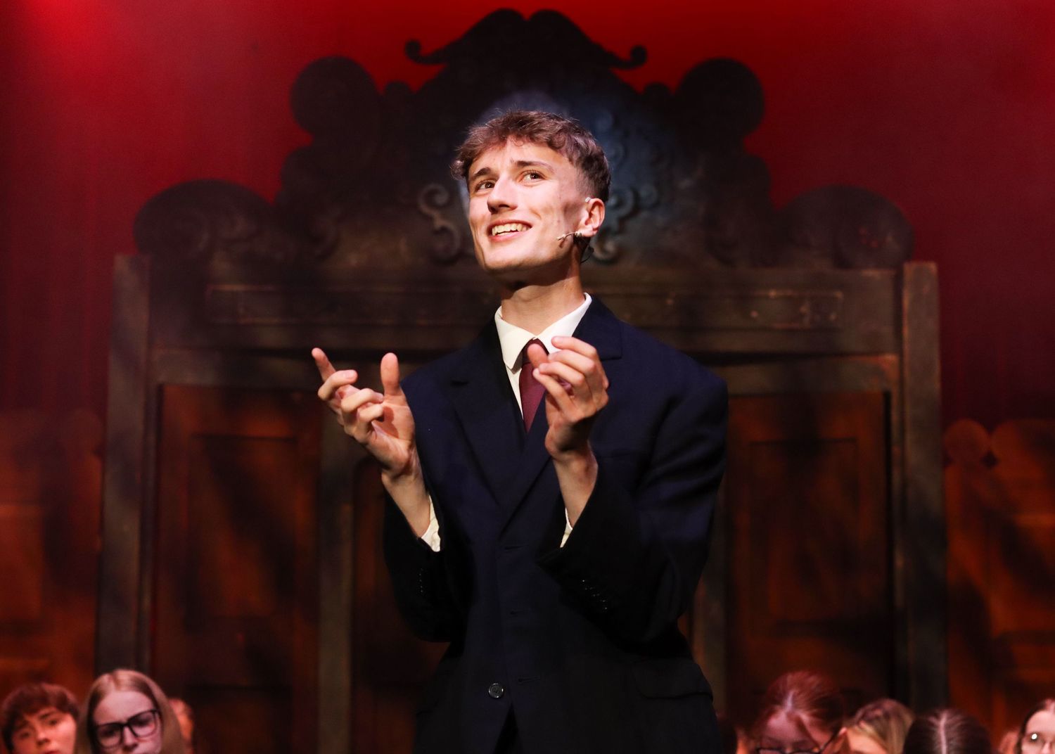Person in dark suit gestures dramatically against red backdrop while performing on ornate wooden stage set.