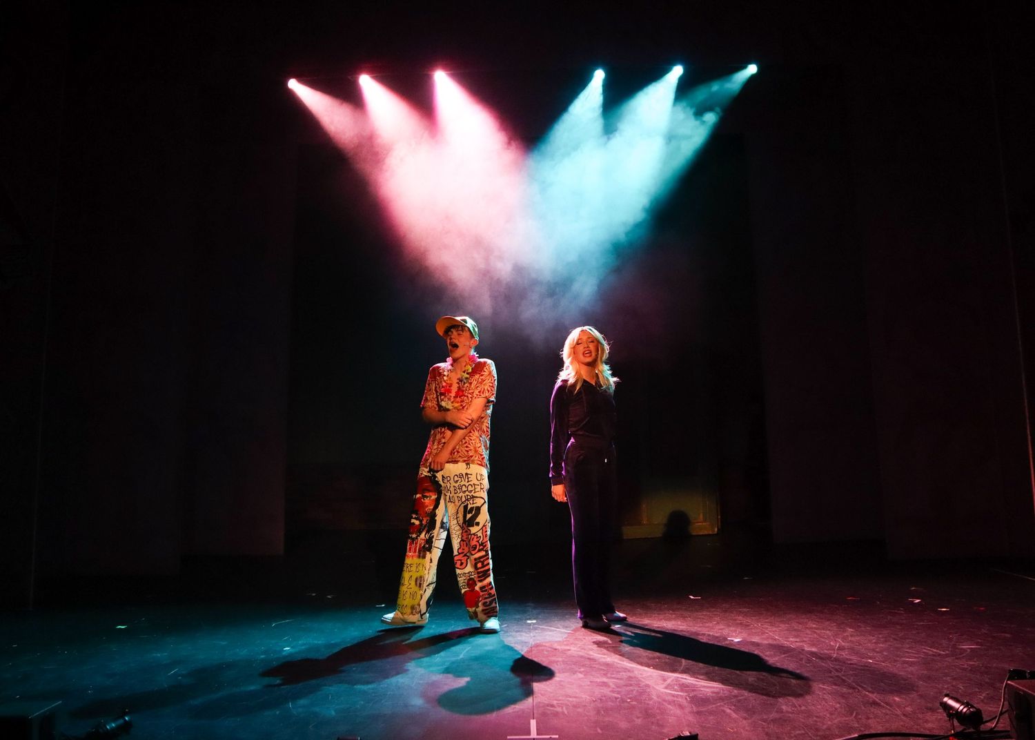 Two performers stand on stage under dramatic pink and blue spotlights during a theatrical performance.
