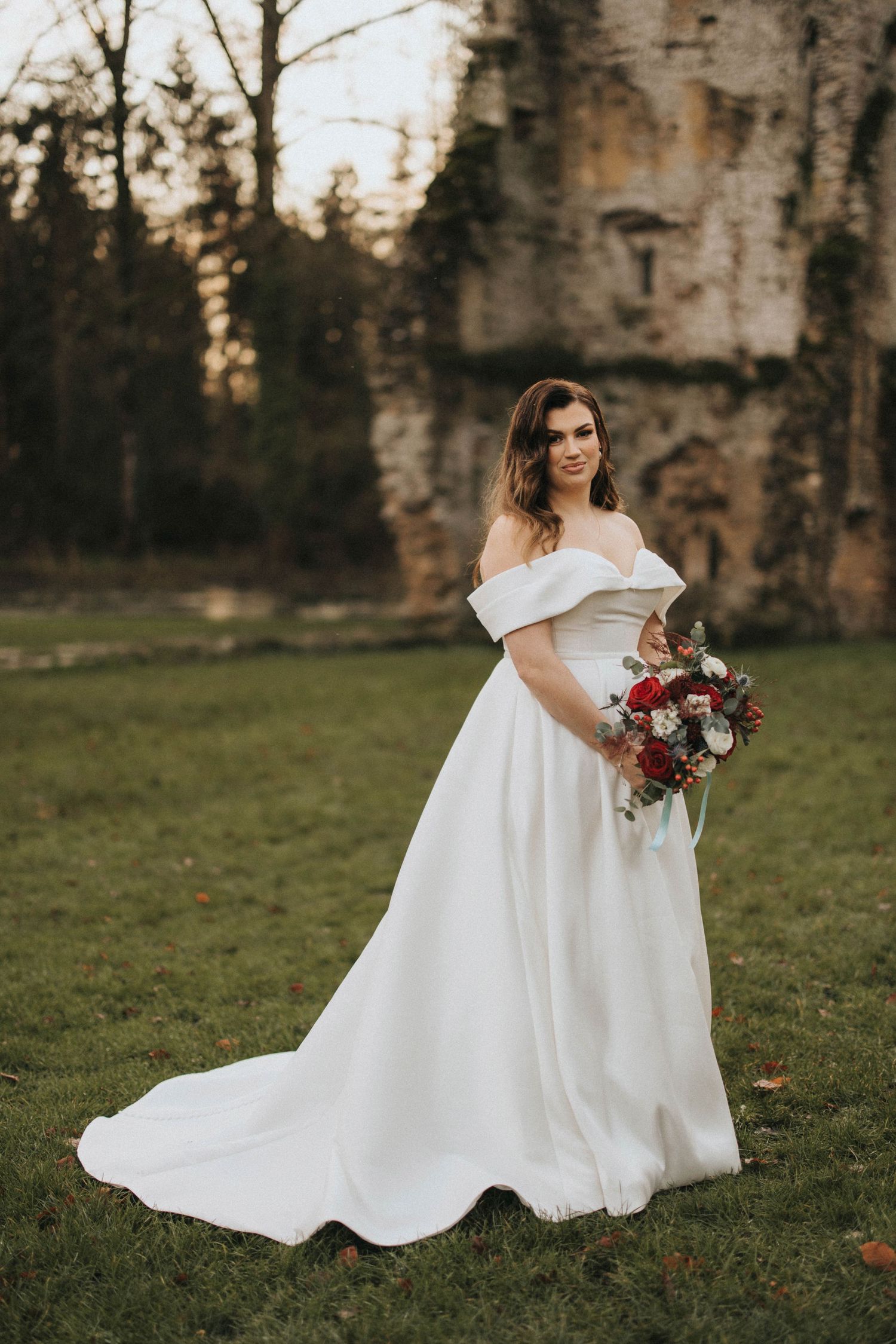 A bride in a flowing white gown twirls on grassy grounds near birch trees during golden hour.