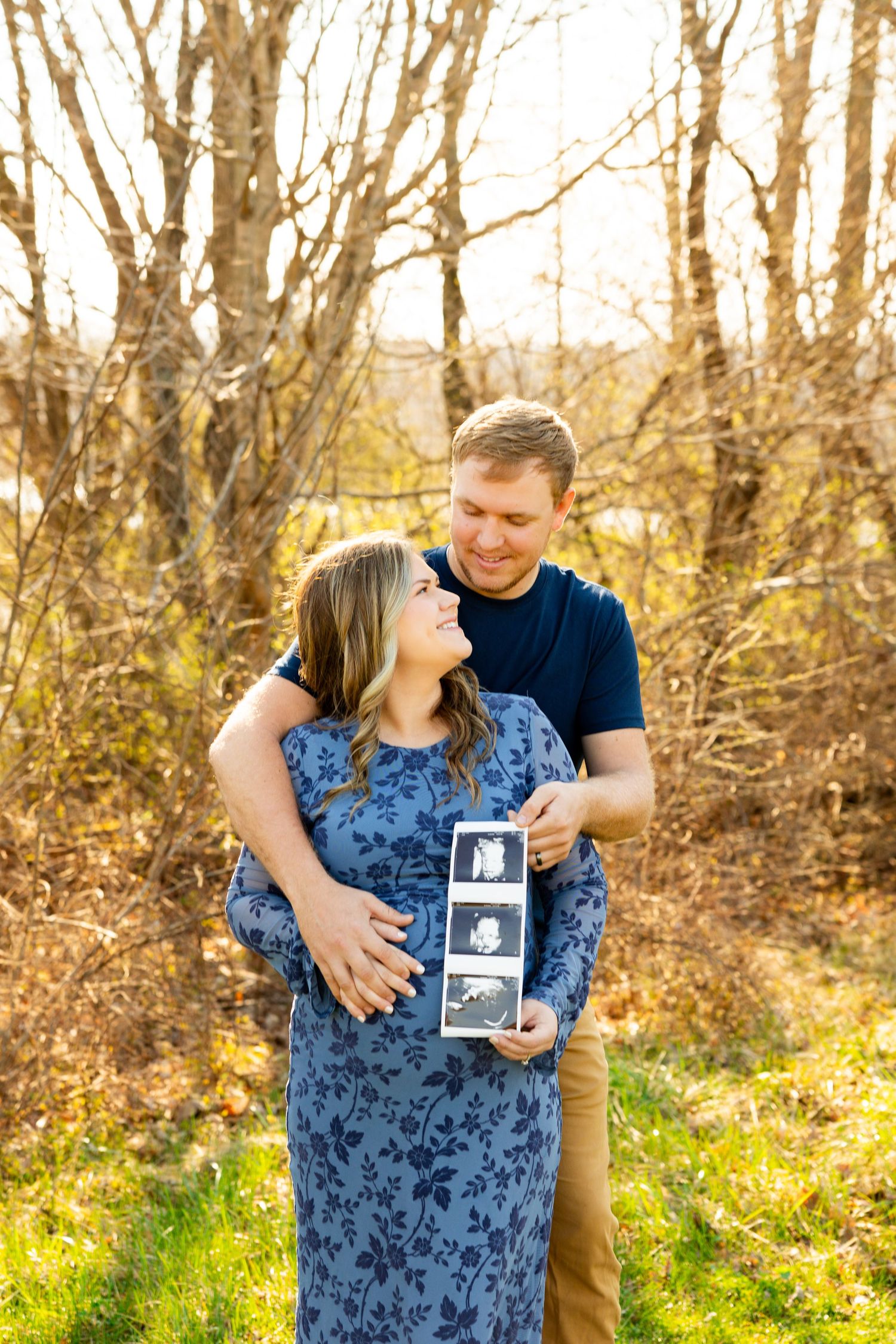 Couple poses outdoors in autumn setting with ultrasound photos during maternity photoshoot.