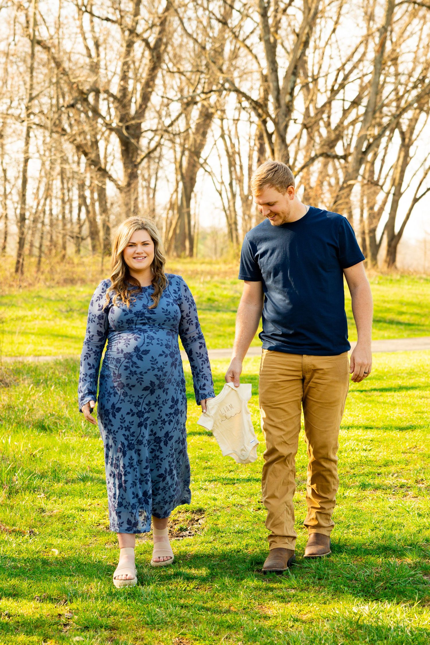 A couple walks together holding hands in a sunny spring meadow with bare trees in the background.