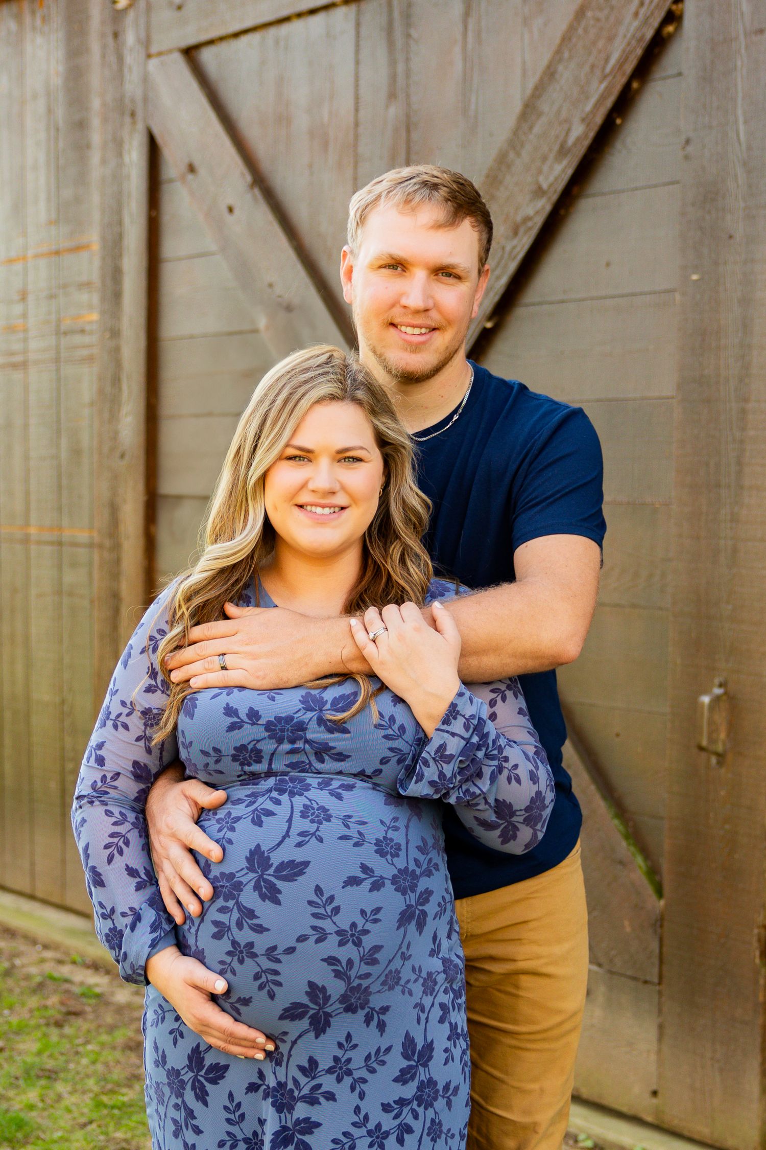 A couple in blue and tan casual clothing pose for outdoor maternity photos against a wooden fence.