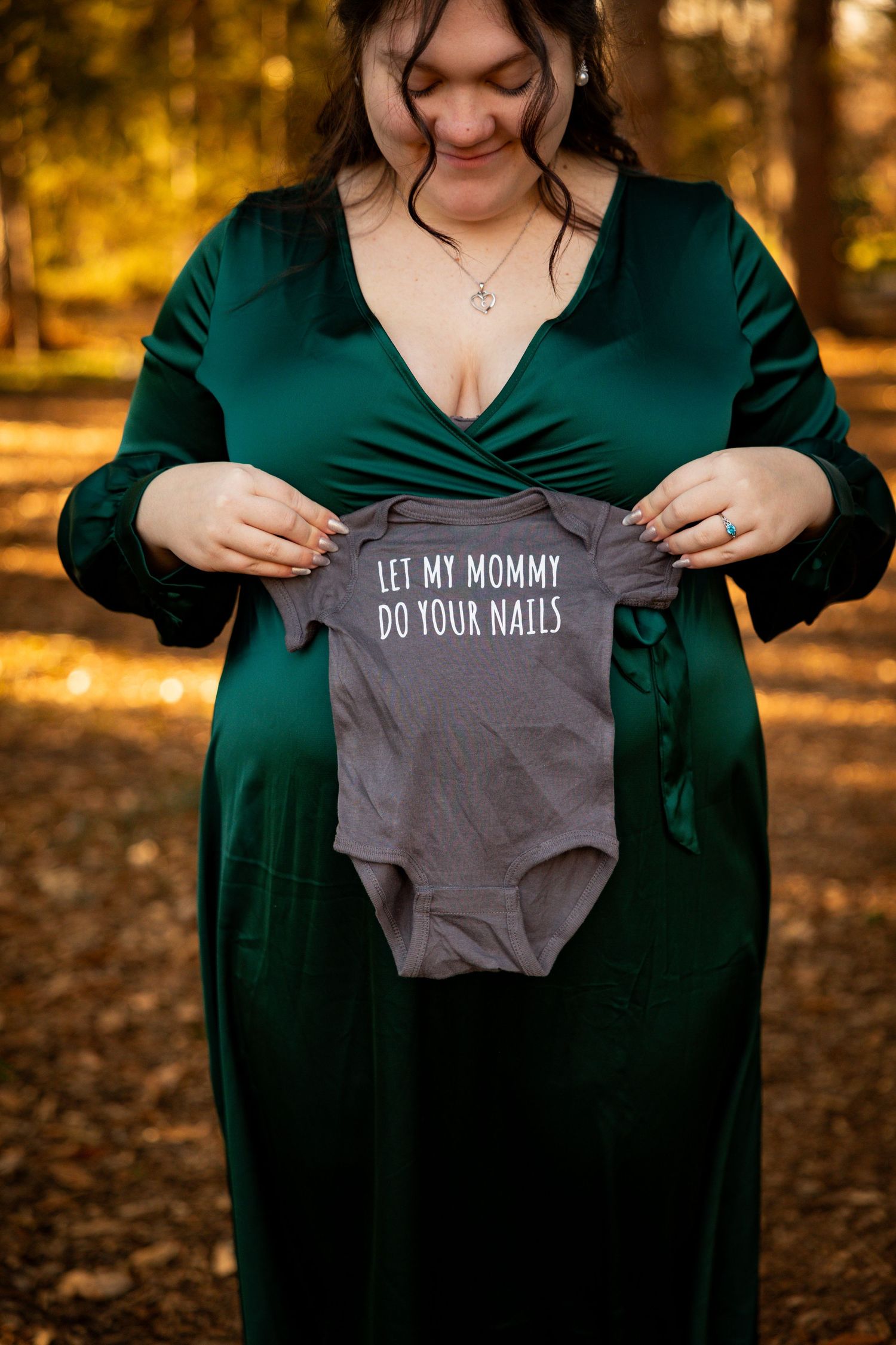 Someone in a dark green dress holds various baby onesies with playful text against an autumn forest backdrop.