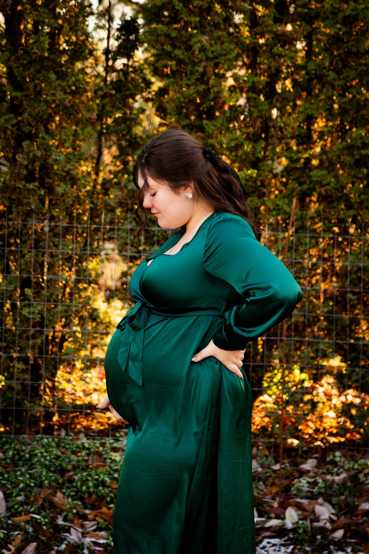 A pregnant woman in an emerald green dress stands against a backdrop of autumn foliage at sunset.