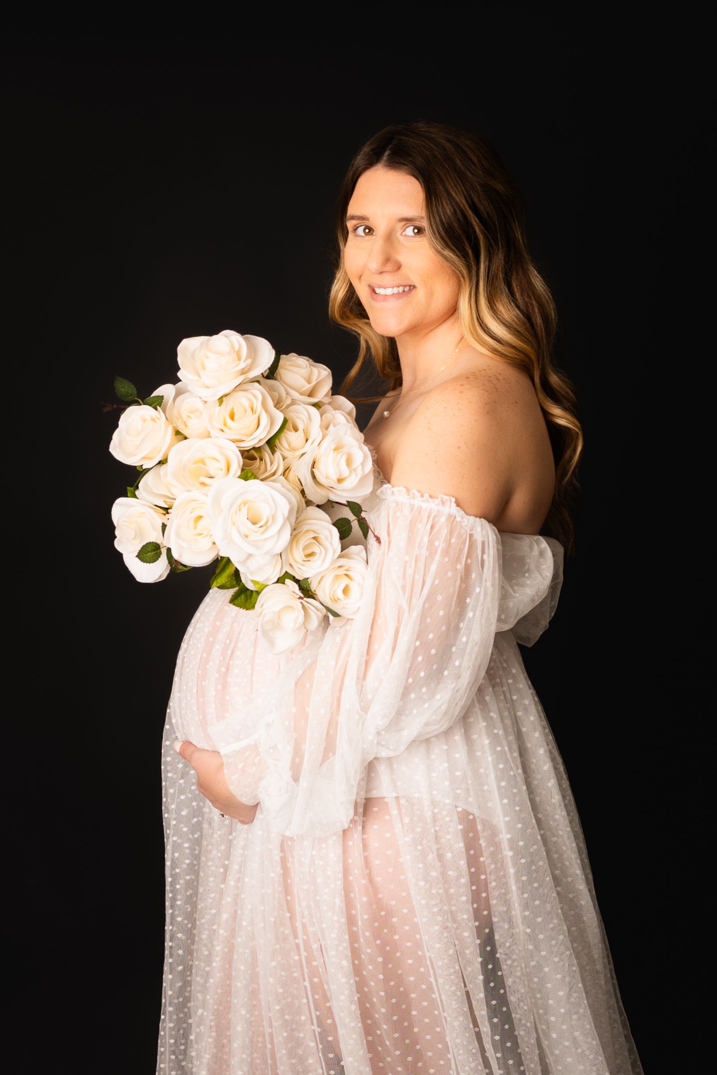 Expecting mother in sheer white polka dot dress holds bouquet of cream roses against dark background.