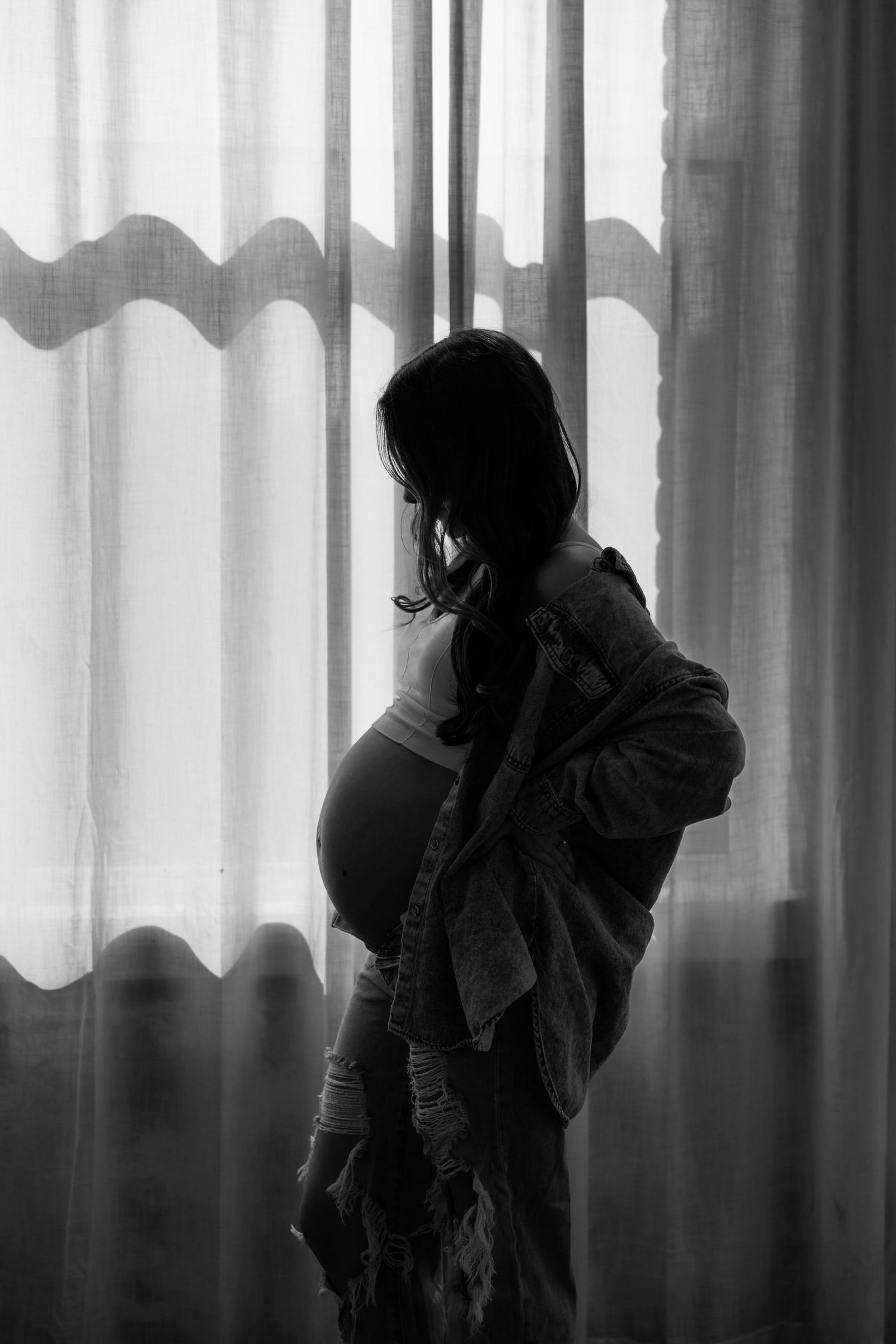 Silhouette of pregnant woman in flowing dress standing by window with sheer curtains in black and white.