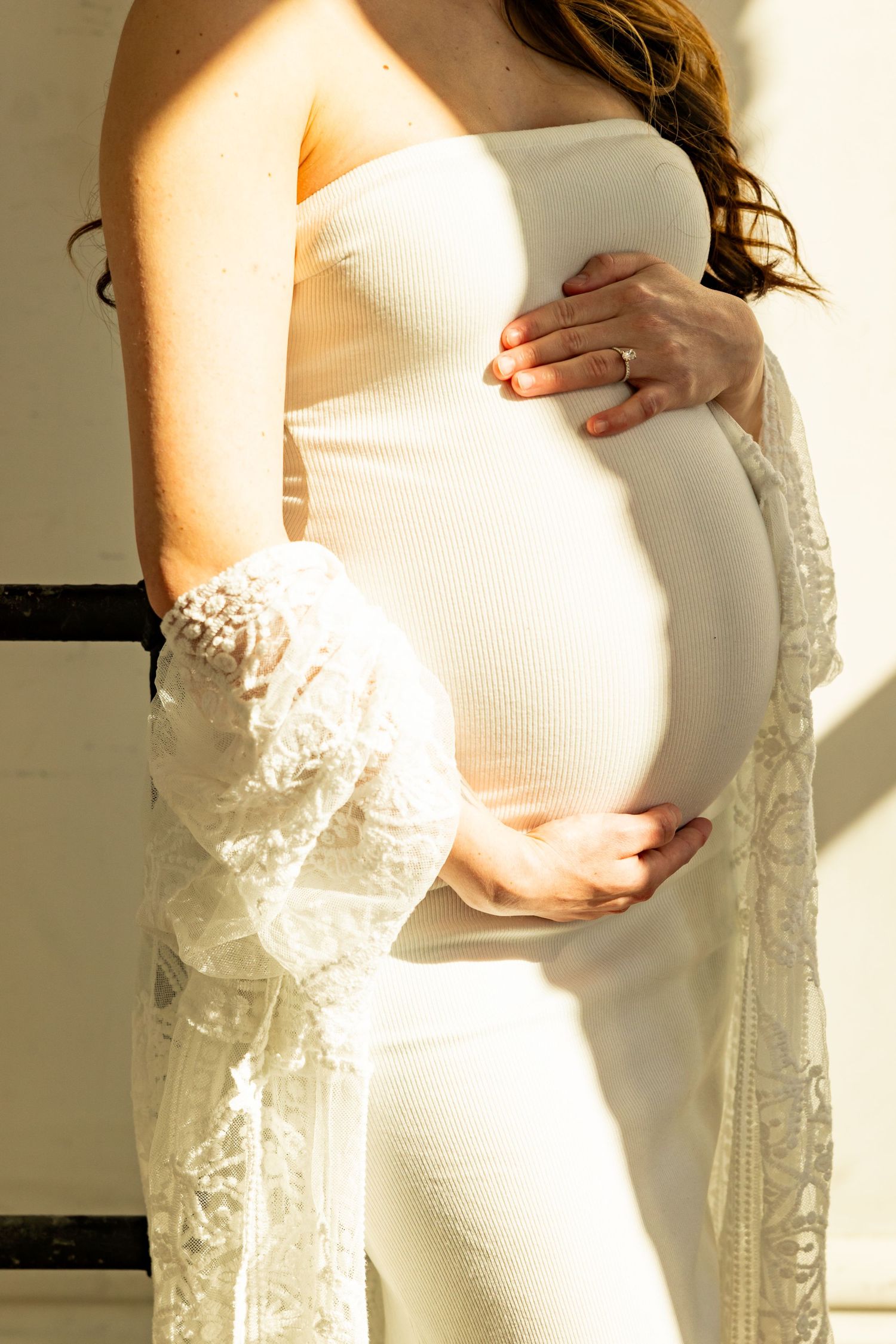 Pregnant woman in white dress cradling baby bump in soft sunlit room with lace details.
