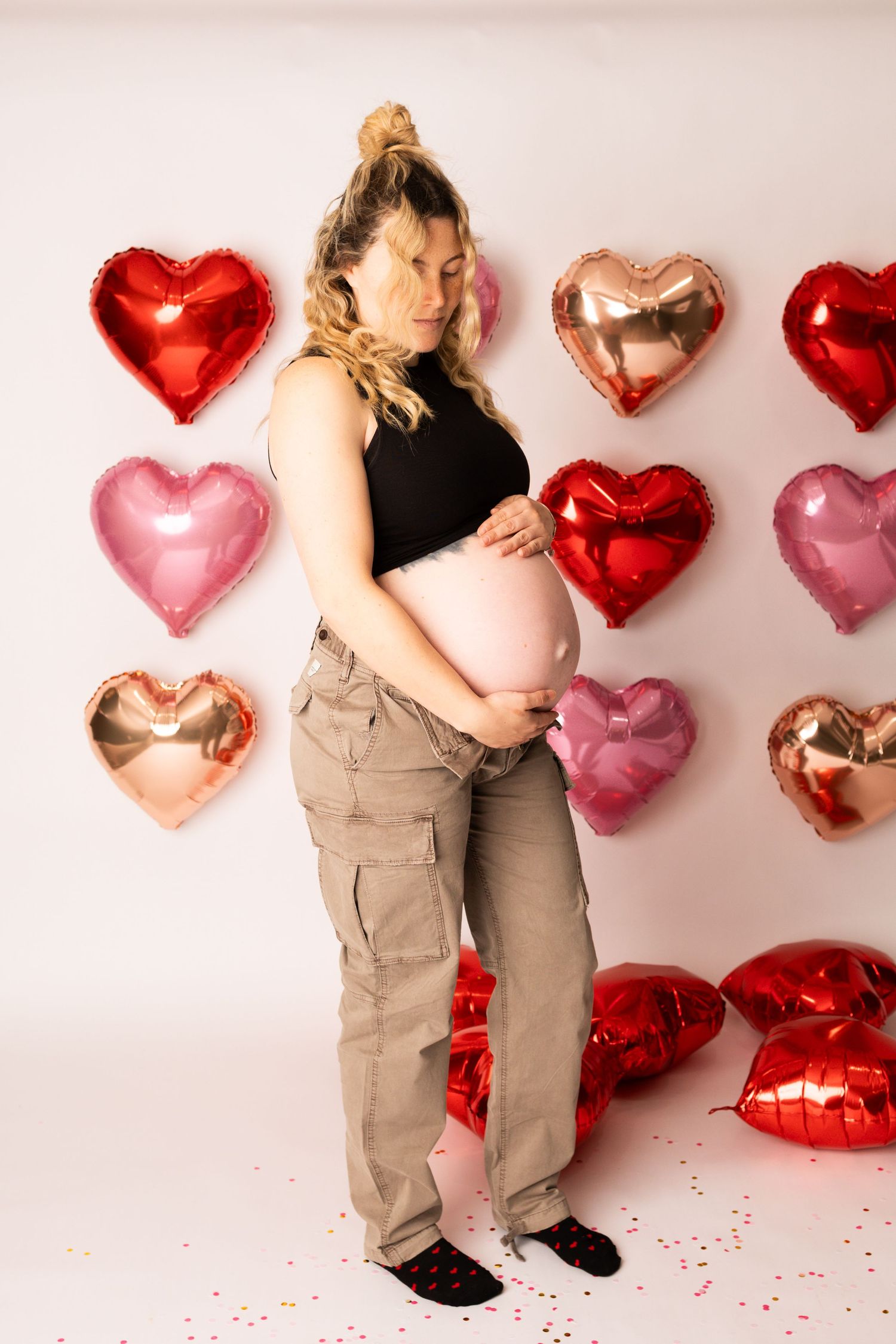 A pregnant person poses in front of pink and red heart-shaped balloons for a Valentine's Day maternity photo.