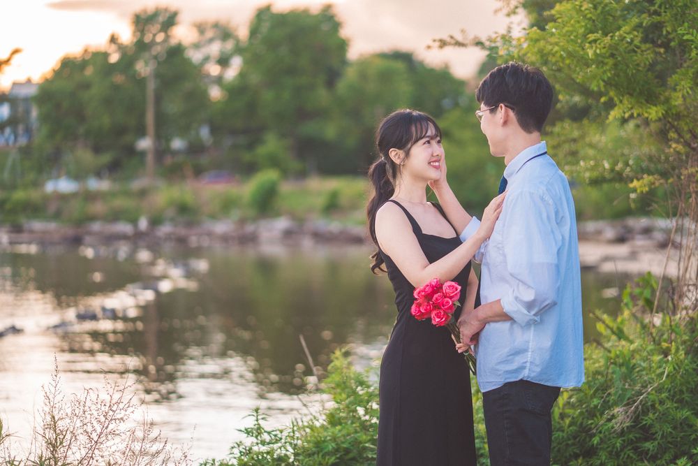 A romantic couple shares a tender moment by a tranquil lake at sunset, with her holding red flowers.