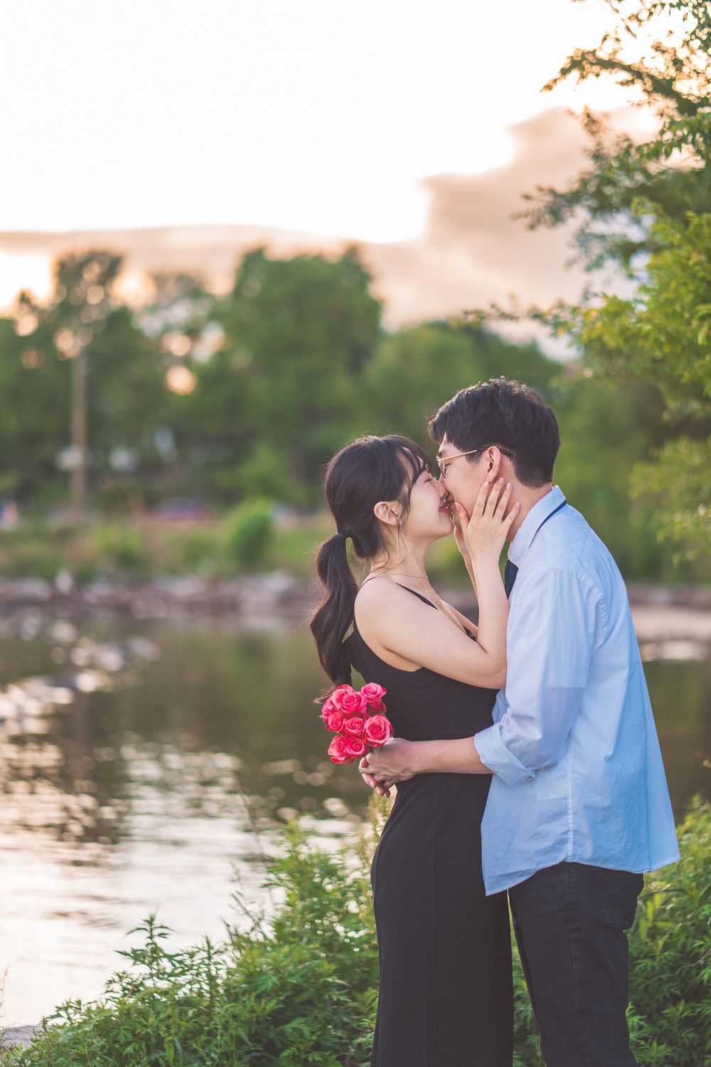 A couple shares a romantic moment at sunset by a lake while holding pink flowers in the garden landscape.