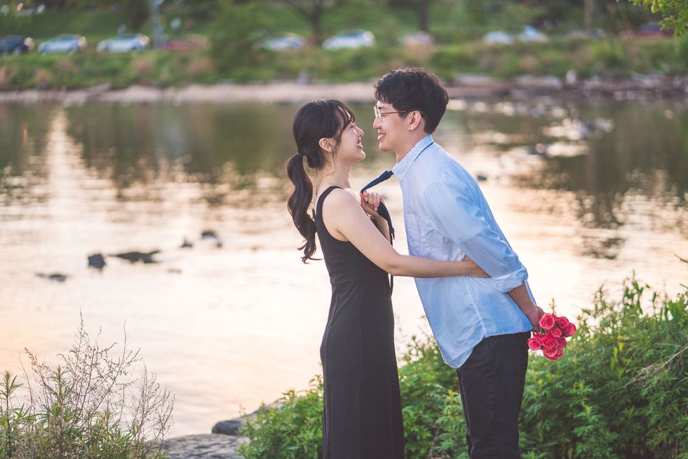 Romantic couple shares a tender moment by a serene lake at sunset as they embrace with red flowers in hand.