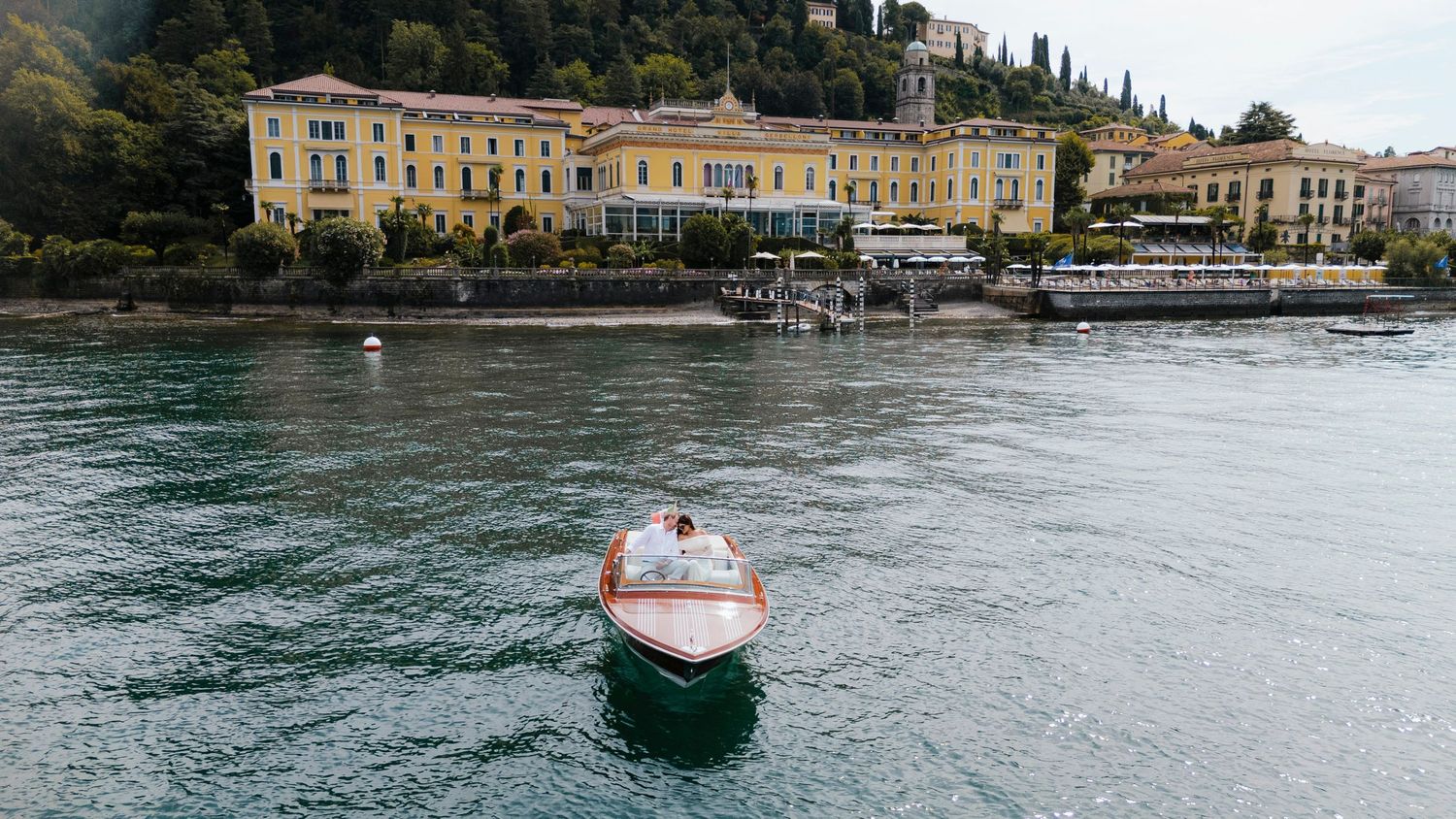 Aerial view of a classic wooden speedboat with blue upholstery cruising through turquoise waters creating white wake.