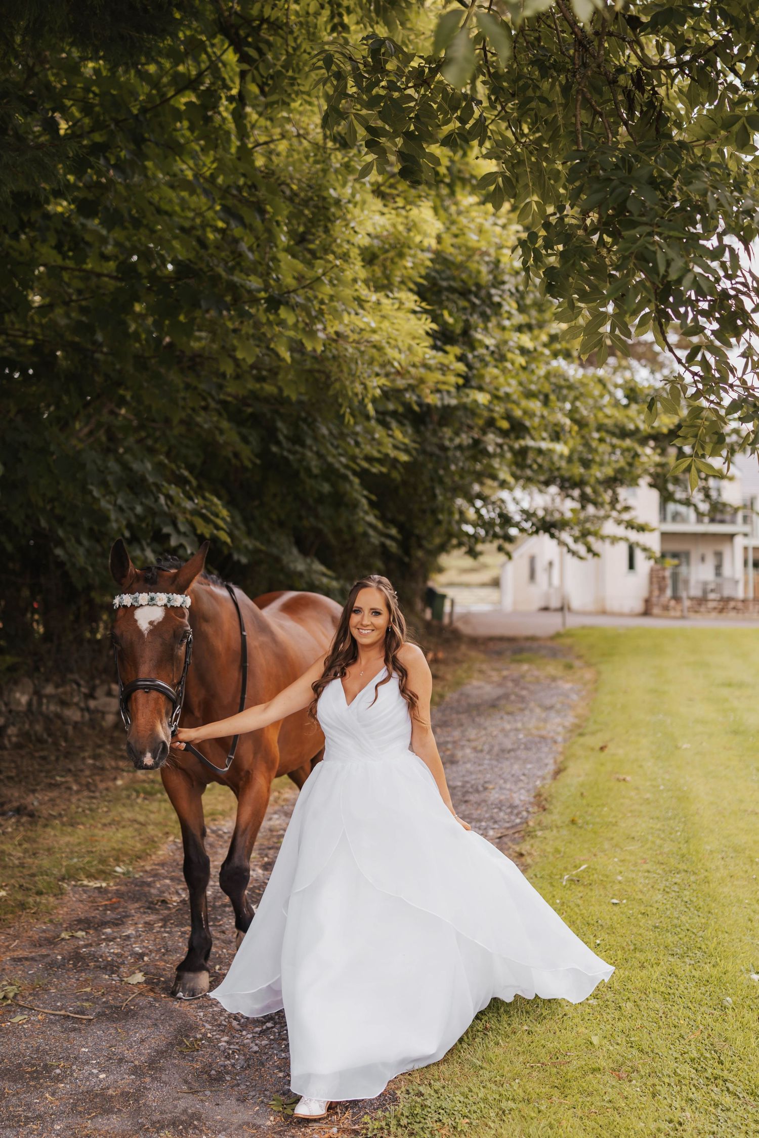 A person in a flowing white wedding dress walks alongside a brown horse on a grassy path under trees.