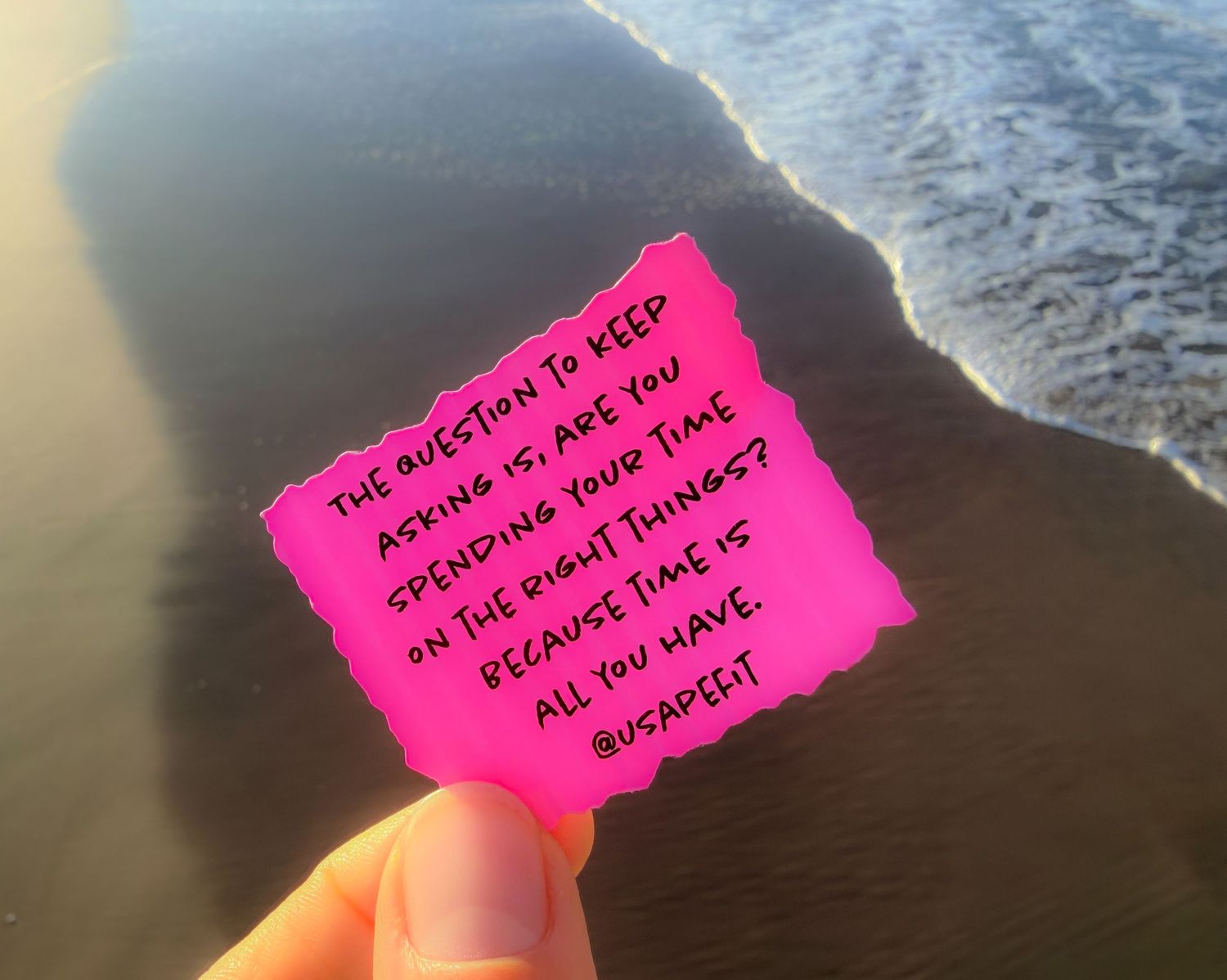 A hand holds a bright pink sticky note against a blurred water background.