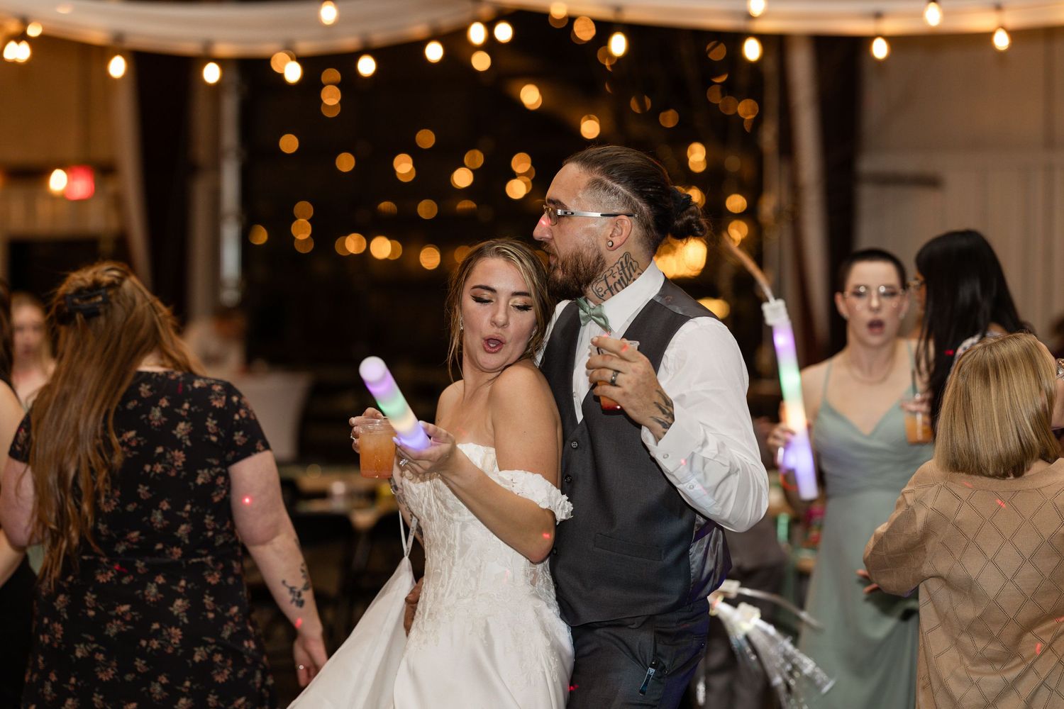Guests dancing and celebrating at an indoor wedding reception with string lights glowing overhead.