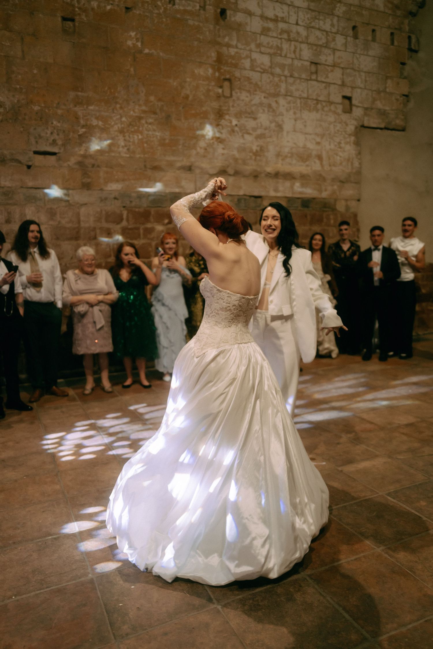 A couple in wedding attire spins and dances together in a rustic brick-walled venue during their first dance.