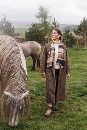 A long-haired pony with flowing mane grazes in a green pasture beside someone in a tweed coat and white scarf.