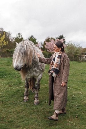A person in a brown coat interacts with a dappled grey horse in a grassy field on an overcast day.