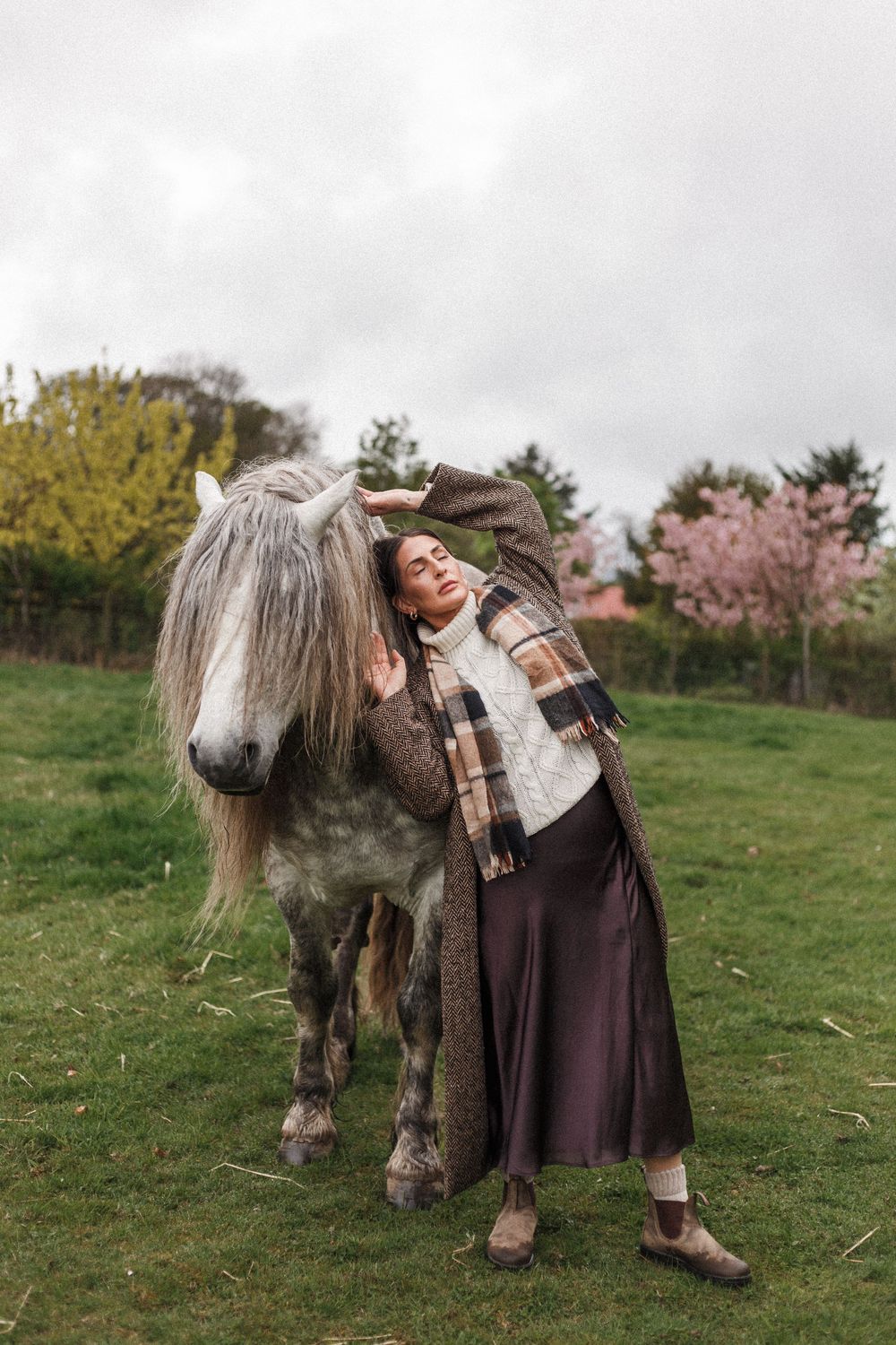 A person in a brown dress and scarf poses affectionately with a small shaggy pony in a grassy field during autumn.